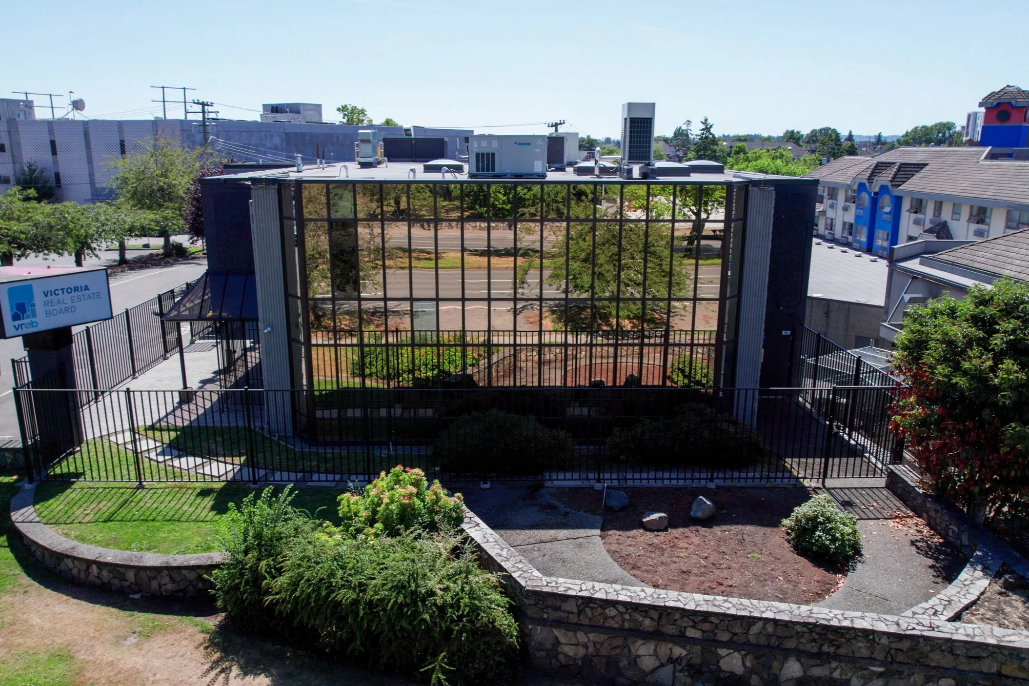 A fenced outdoor area with a large glass pane structure, surrounded by greenery, trees, and buildings, with a sign reading 'Victoria Real Estate Board' on the left.