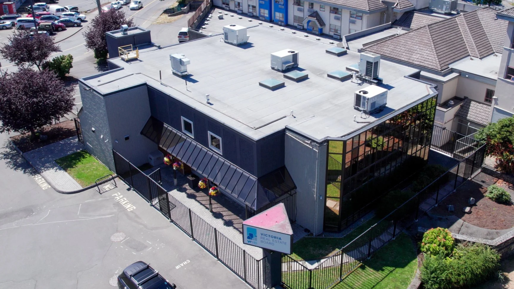 Aerial view of a commercial building with a flat roof, surrounded by a parking lot and fencing, with trees and neighboring buildings nearby.