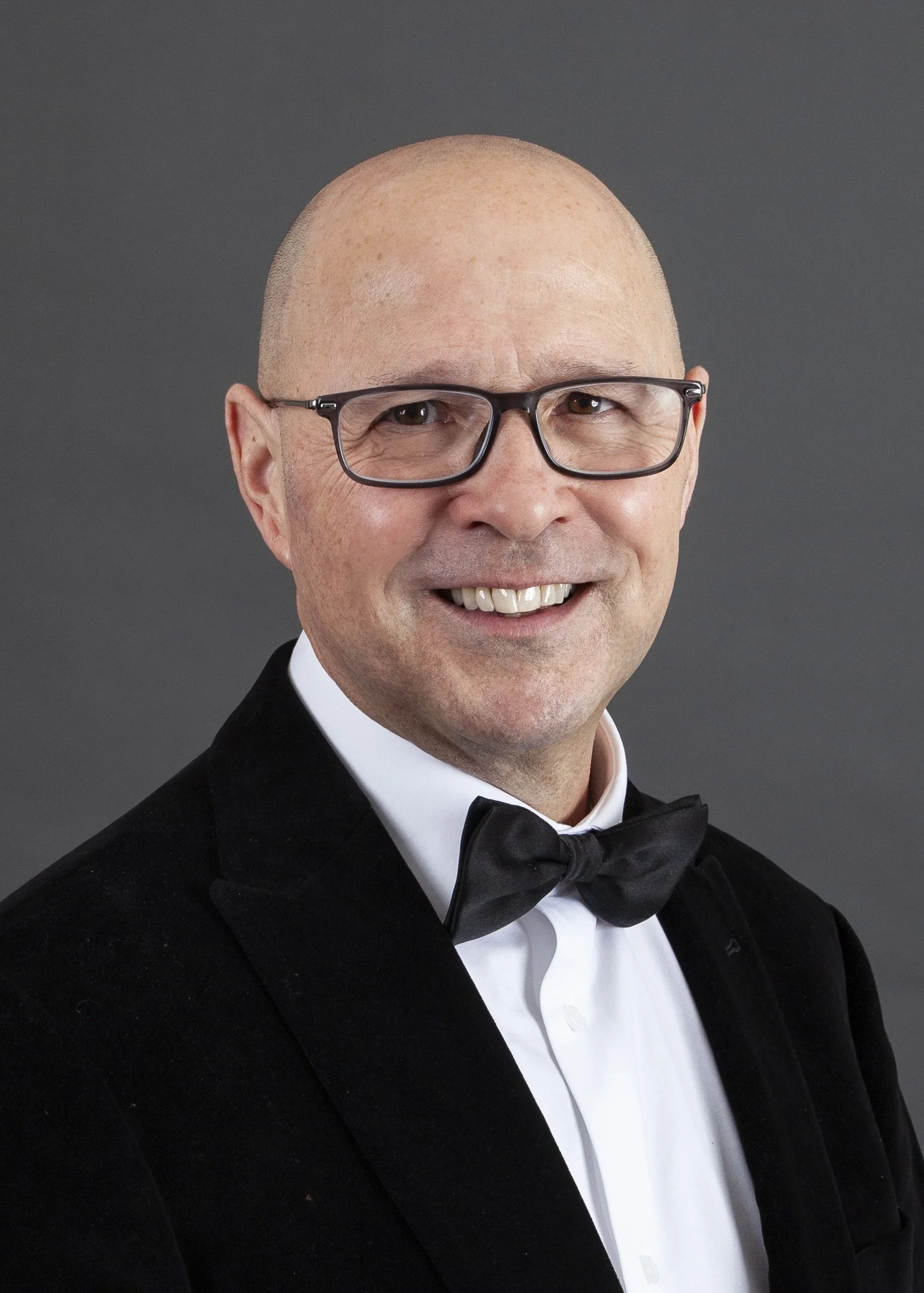 Headshot of a man with glasses, smiling, wearing a white shirt and a dark blazer against a plain background.