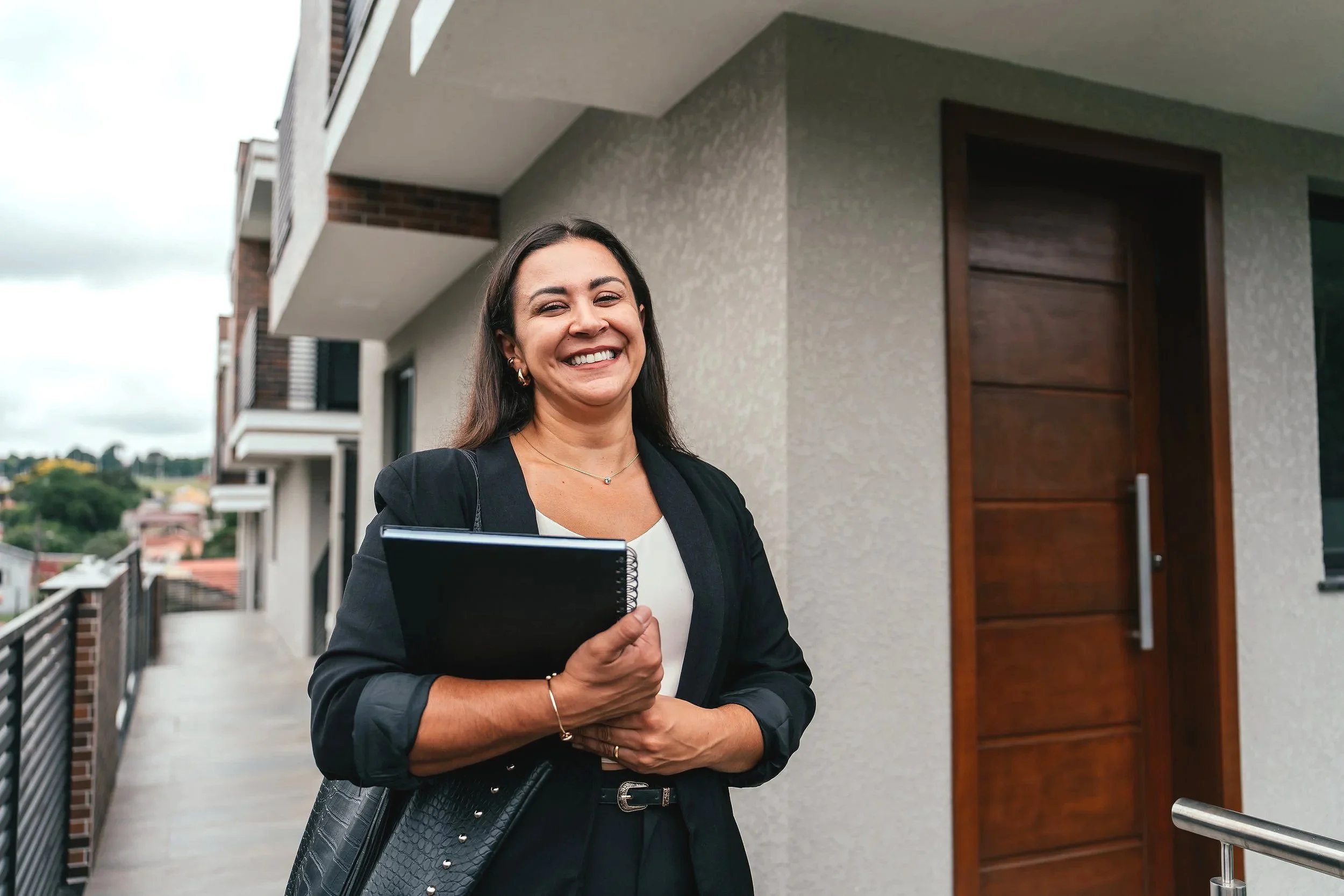 A smiling woman standing outside a modern condo building, holding a folder and a handbag.