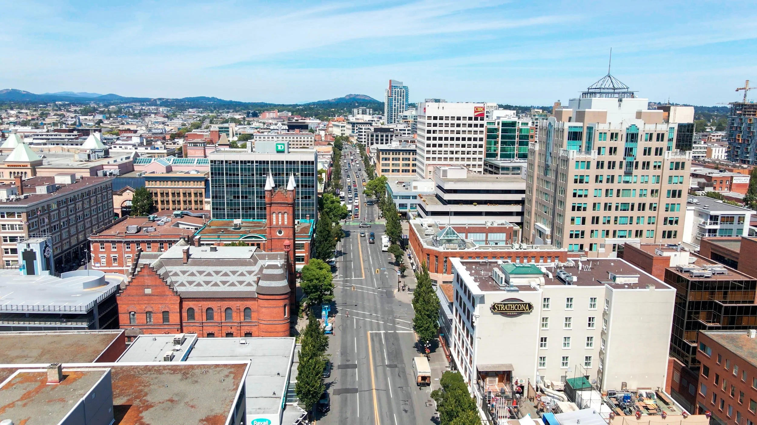Aerial view of a city street with buildings, trees, and cars under a blue sky.