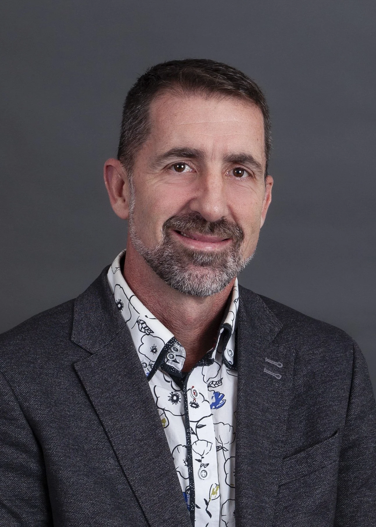 Professional headshot of a middle-aged man with short dark hair, a beard, and mustache, wearing a dark blazer over a white dress shirt with a colorful abstract pattern, against a plain gray background.