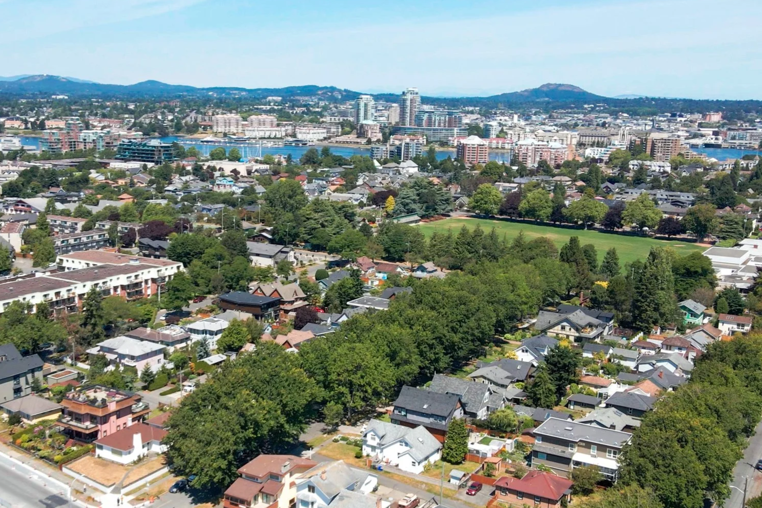 Aerial view of a city with a mix of residential houses, a large green park, water body, and high-rise buildings in the background, with hills in the distance.