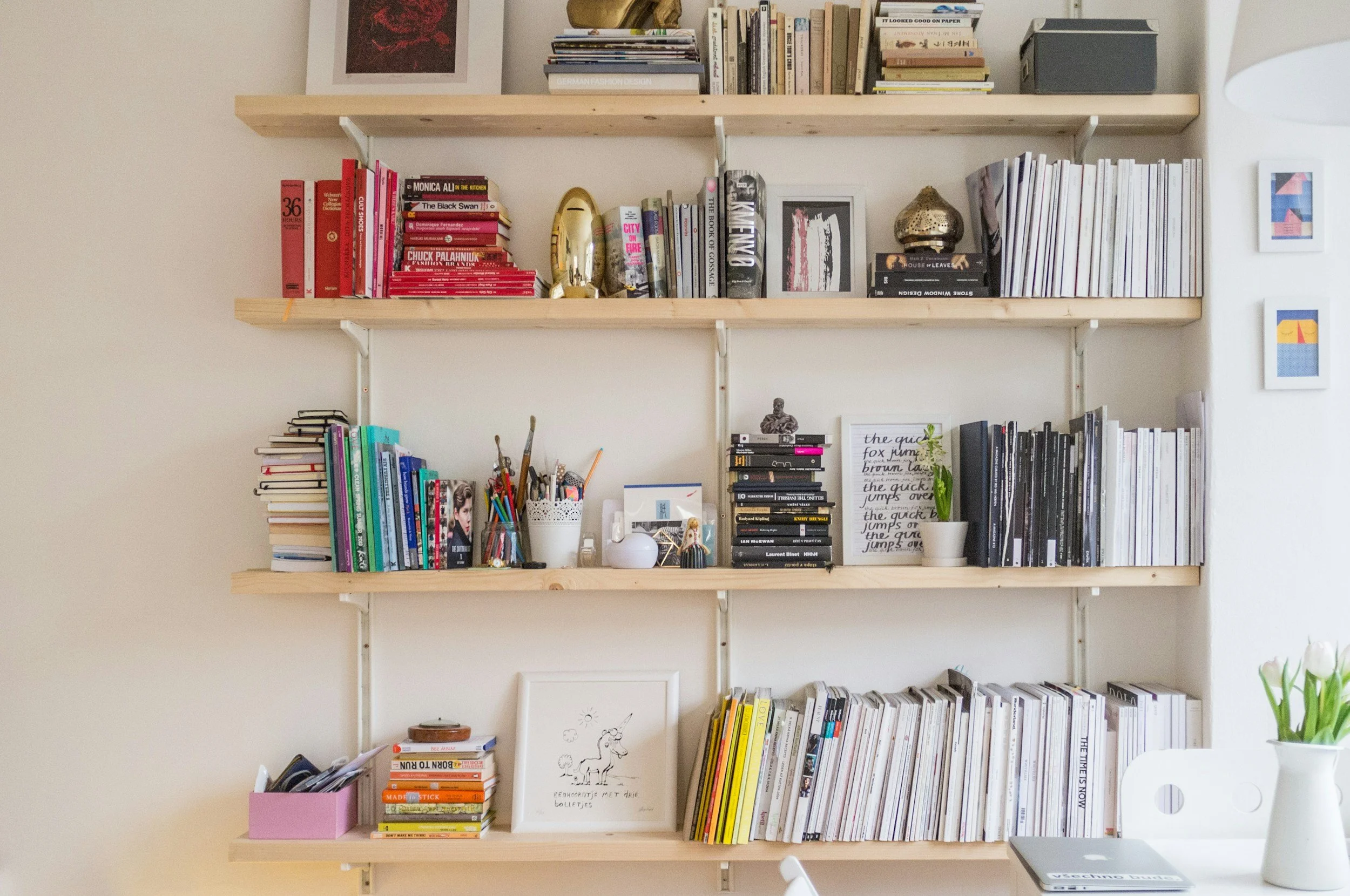 A bookshelf filled with various books, decorative items, and framed artwork against a white wall, with a white desk and vase of tulips nearby.