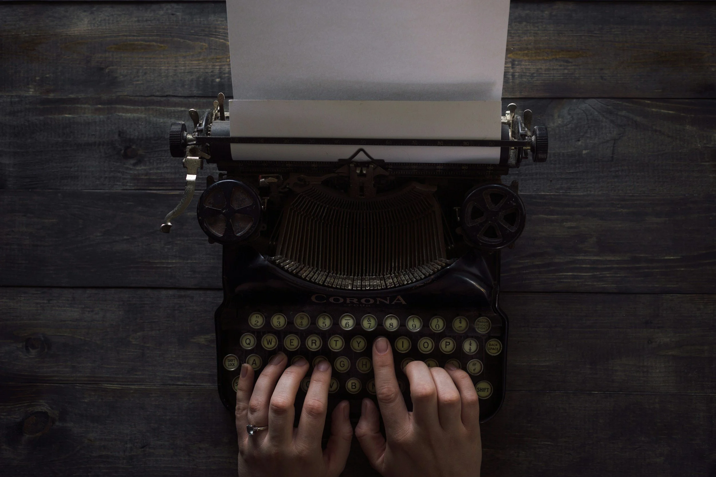 A vintage black typewriter with a sheet of paper inserted, on a dark wooden surface, with hands typing on the keys.