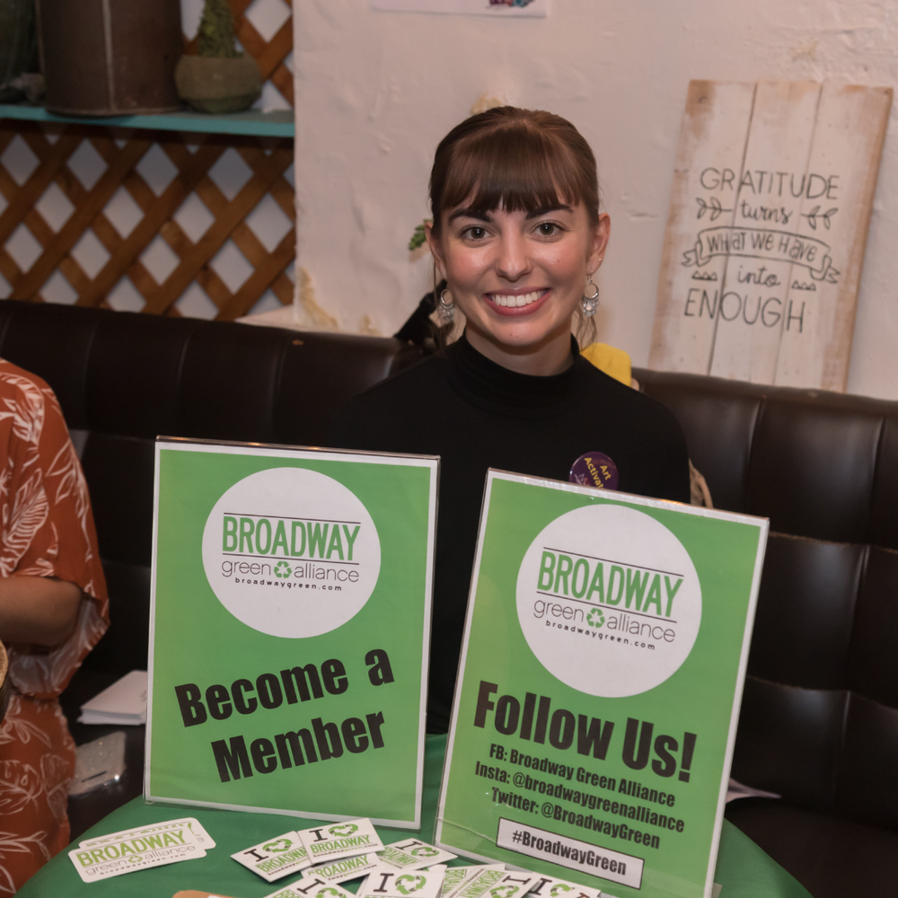 A woman with brown hair and bangs smiling sitting at a table with green promotional signs for Broadway Green Alliance, encouraging people to become members and follow on social media.