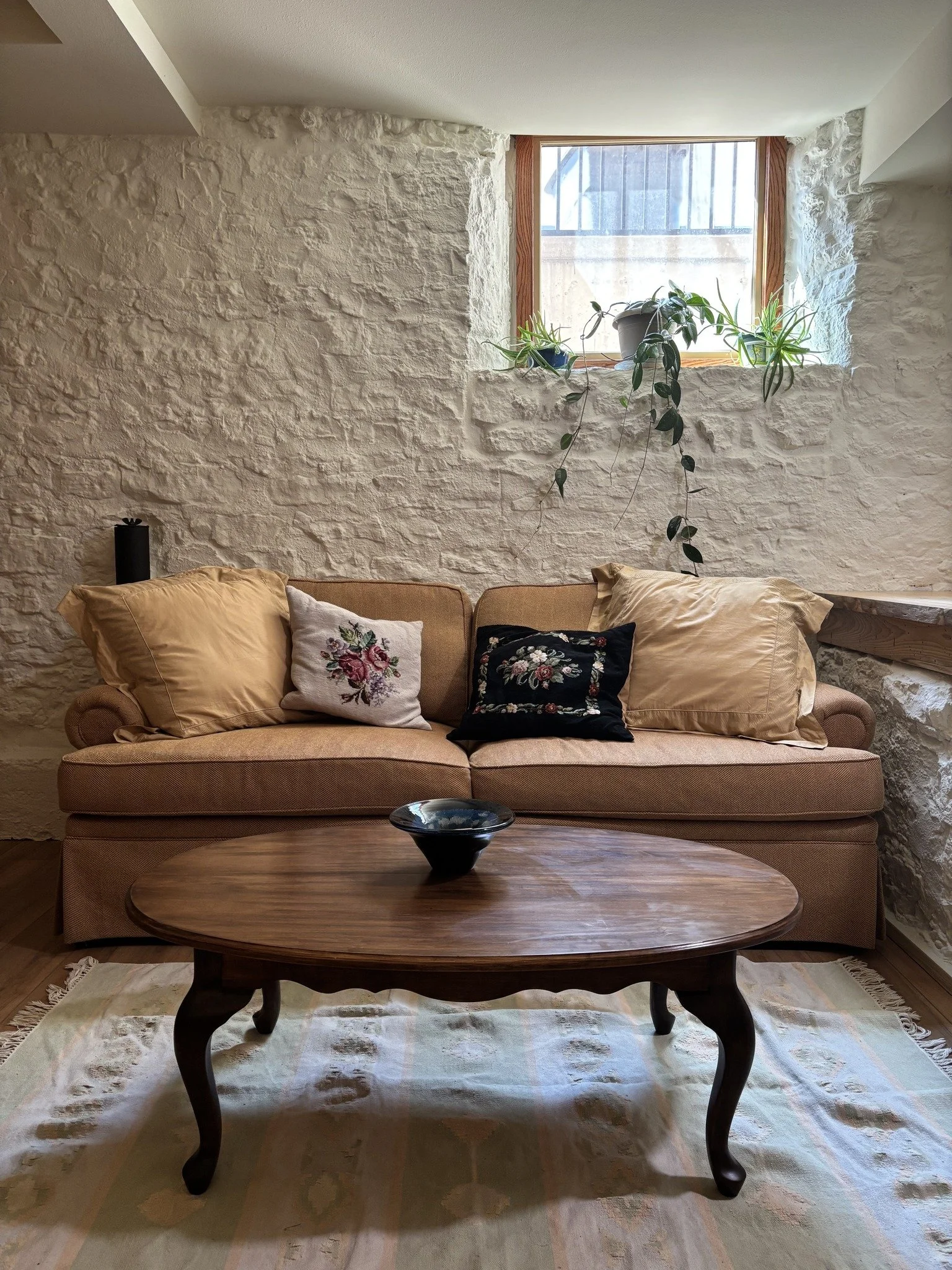 Living room with a beige couch against a white, textured stone wall. There are three pillows on the couch, two beige and one black with embroidery. A wooden coffee table in front of the couch holds a black bowl. A small window above the couch has a wooden frame, with potted plants on the windowsill. A light-colored rug with patterns covers the floor.
