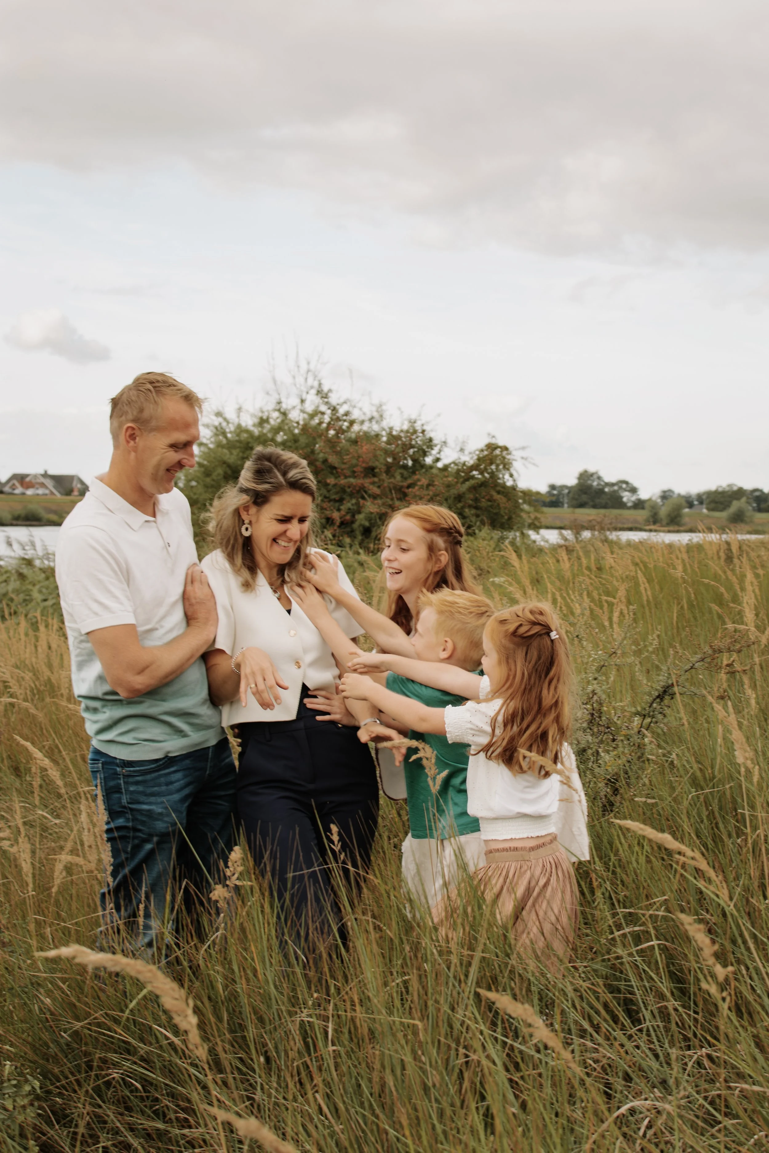 Een gezin met twee volwassenen en vier kinderen staat in een veld met hoog gras en het lijkt op een natuurrijke omgeving buiten, waarschijnlijk in de natuur.