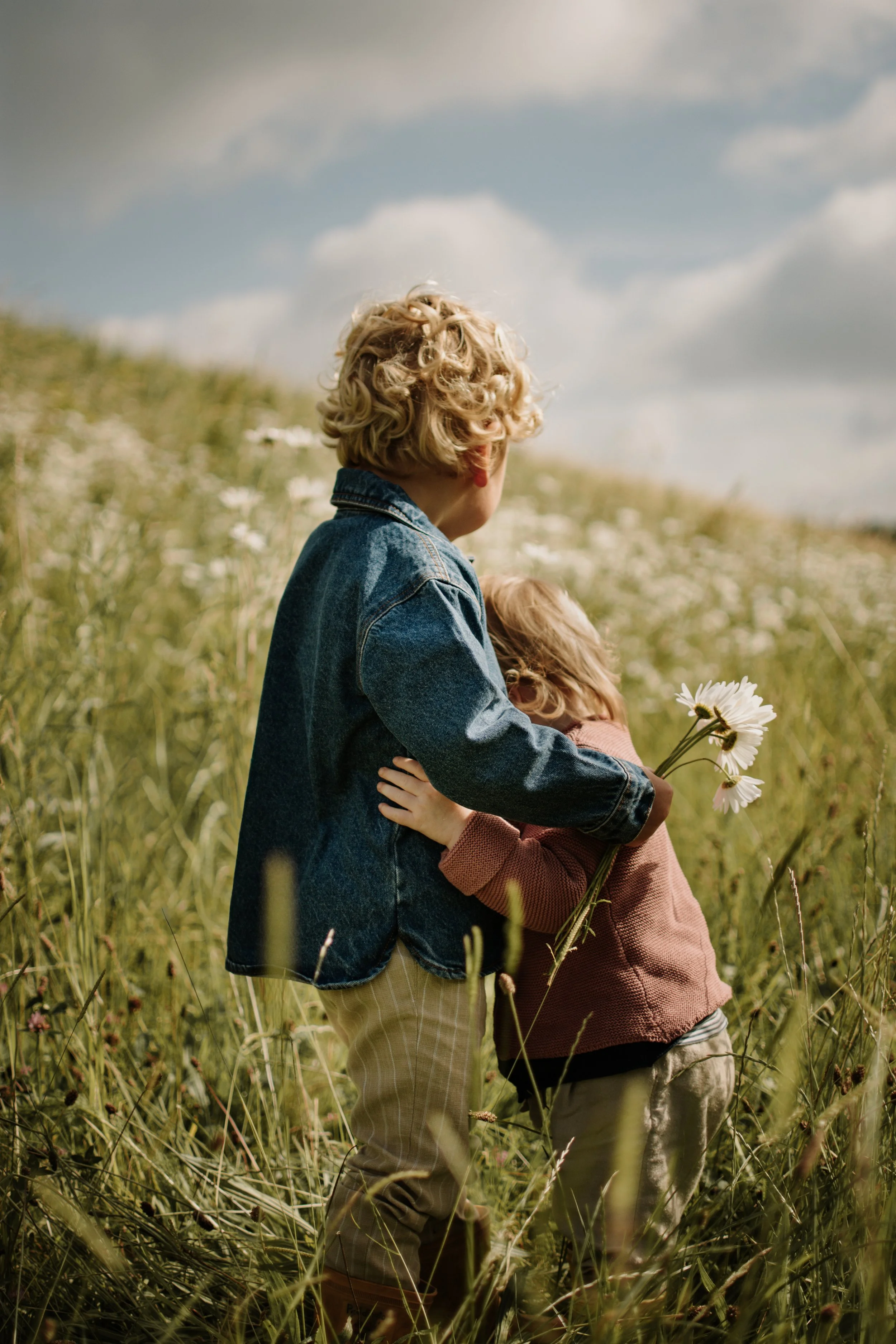 Twee kinderen staan in een veld vol gras en bloemen, met een heldere blauwe lucht en wolken op de achtergrond, één kind houdt bloemen vast.