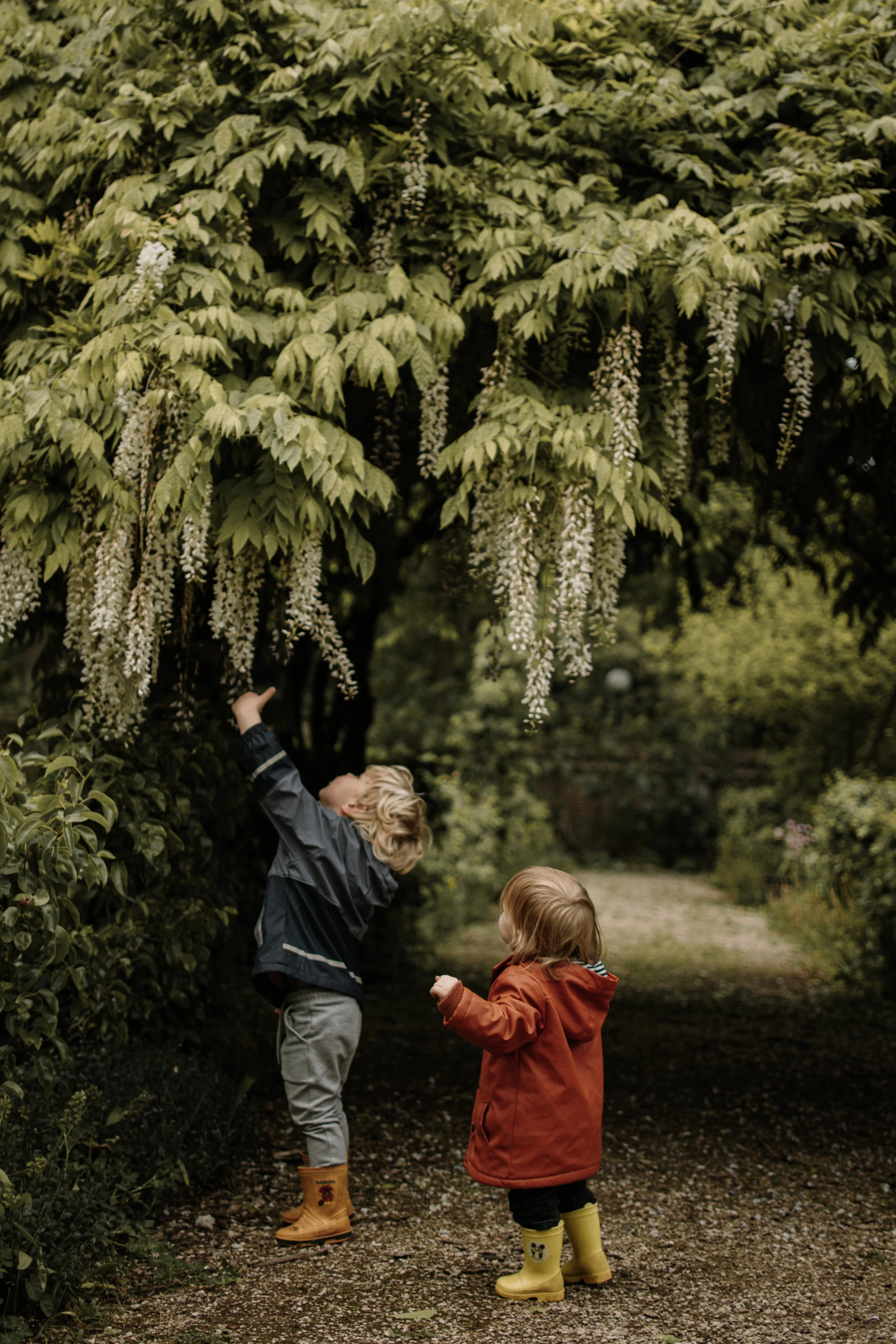 Twee jonge kinderen die in een tuin onder een bloeiende boom staan, met rubberen laarzen aan.