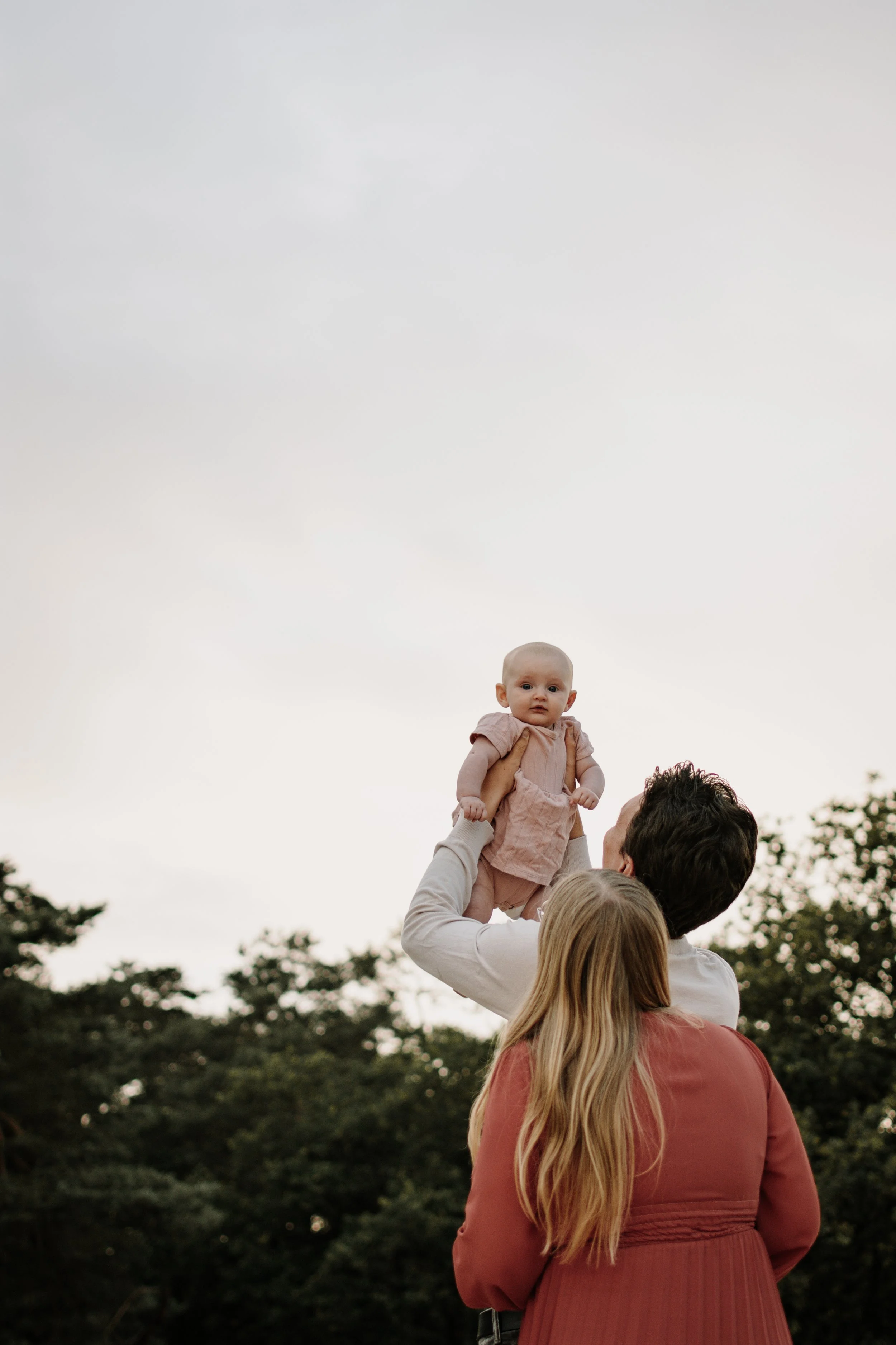 Een persoon met lang blond haar in een rokje en jas, die een baby vasthoudt die op een schouder ligt, terwijl een man het kind omhoog tilt, tijdens een picknick in de natuur.