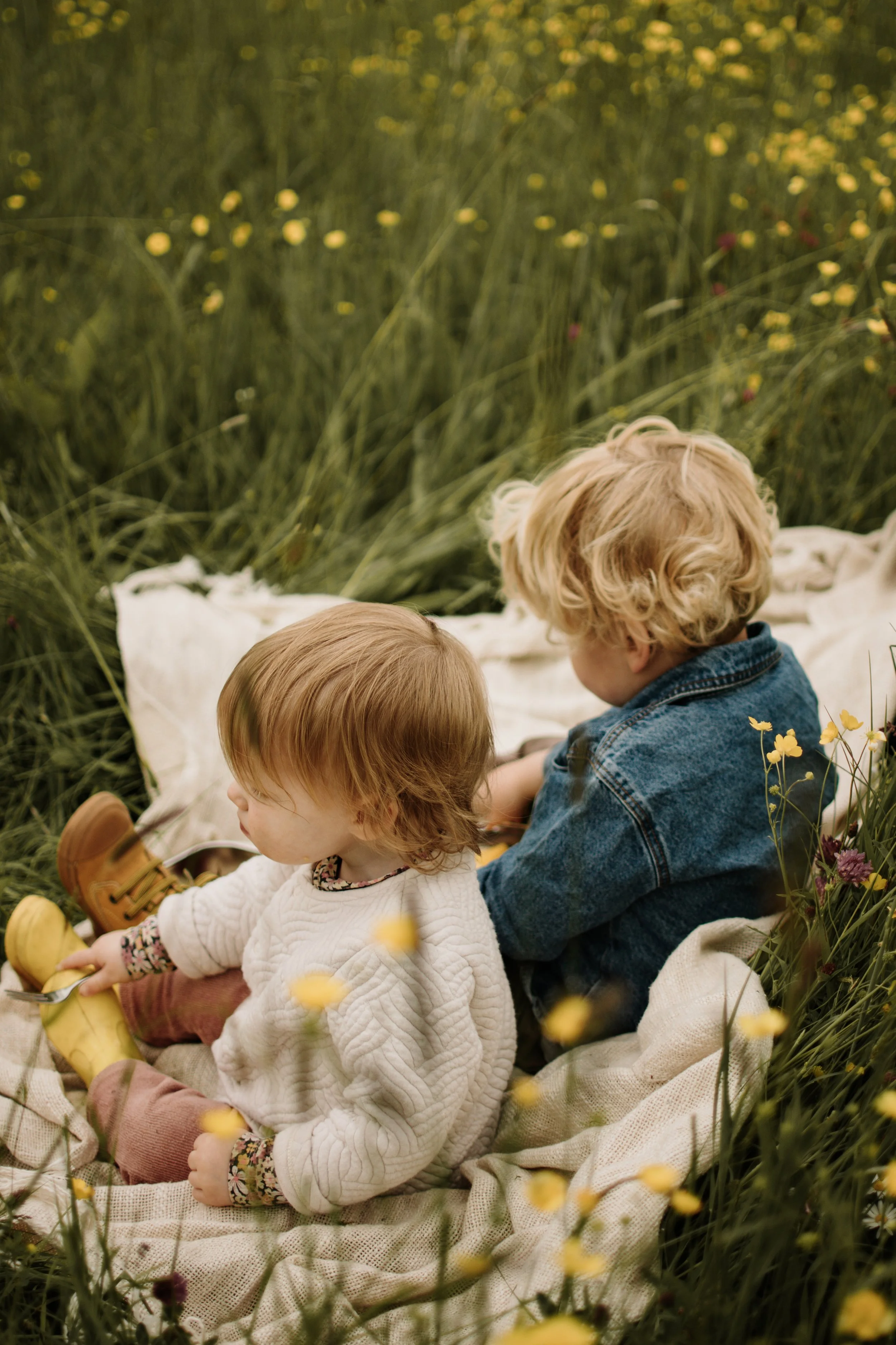 Twee kinderen zitten op een blanket in een veld met gele bloemen, een meisje met een witte trui en een jongen met een denim jas.