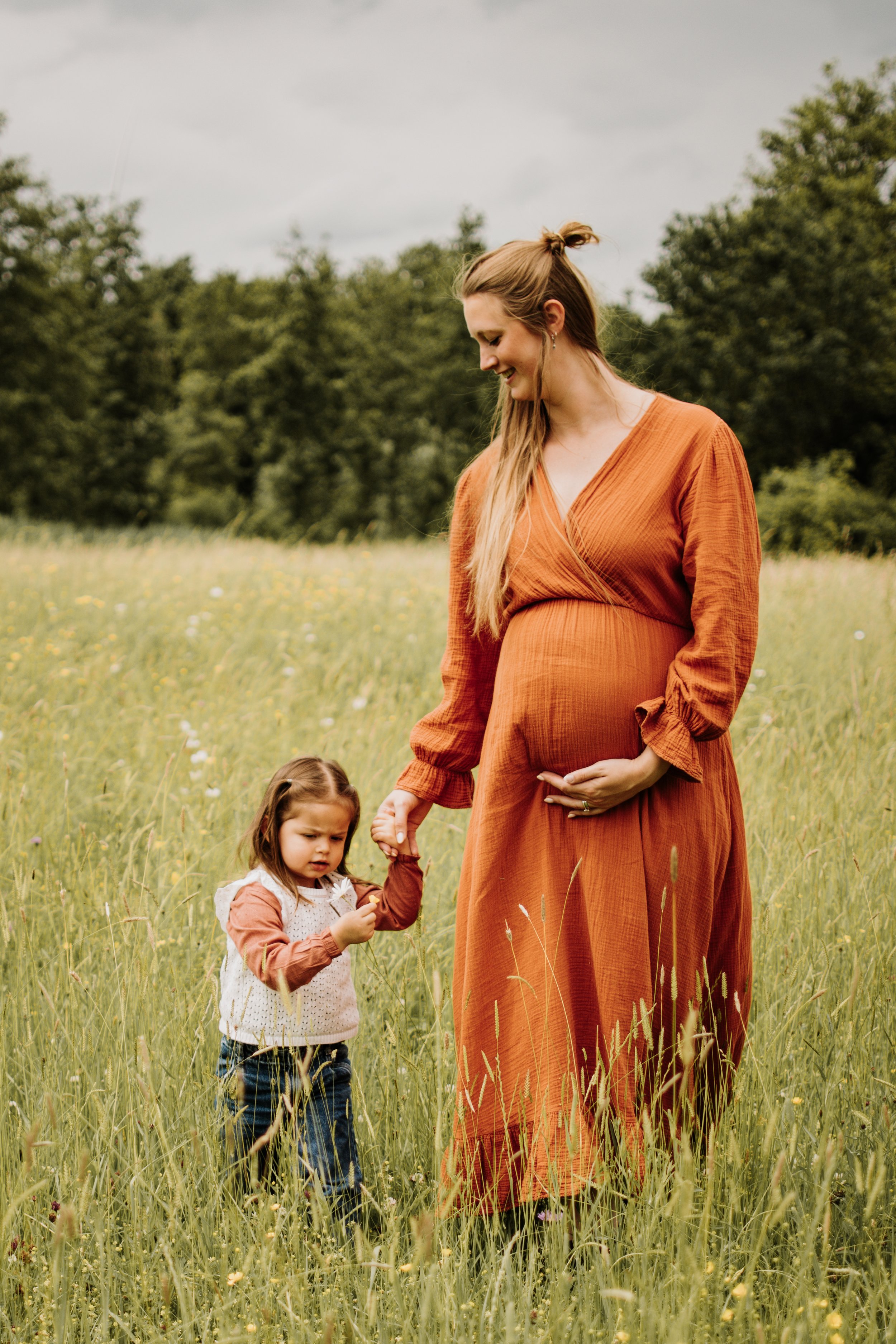Vrouw en twee jonge meisjes in een veld met groen gras en bloemen, met bomen en een bewolkte lucht op de achtergrond.