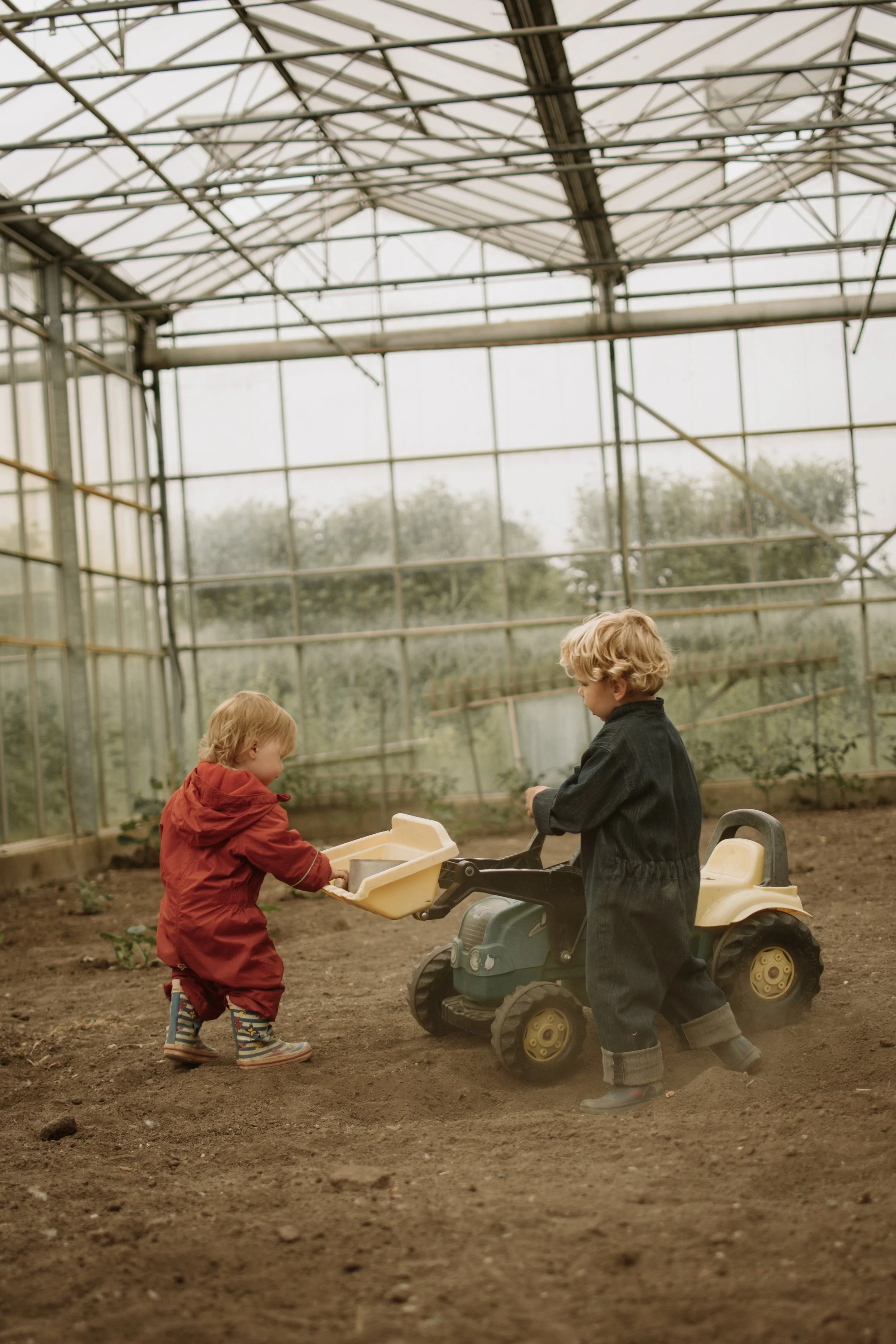 Twee jonge kinderen spelen samen in een grote serre met een metalen structuur en grote ramen. Eén kind zit in een kleine speelgoedauto, terwijl de ander een zandbak of soortgelijke container vasthoudt. Ze dragen regenwaterdichte kleding en rubberen laarzen.