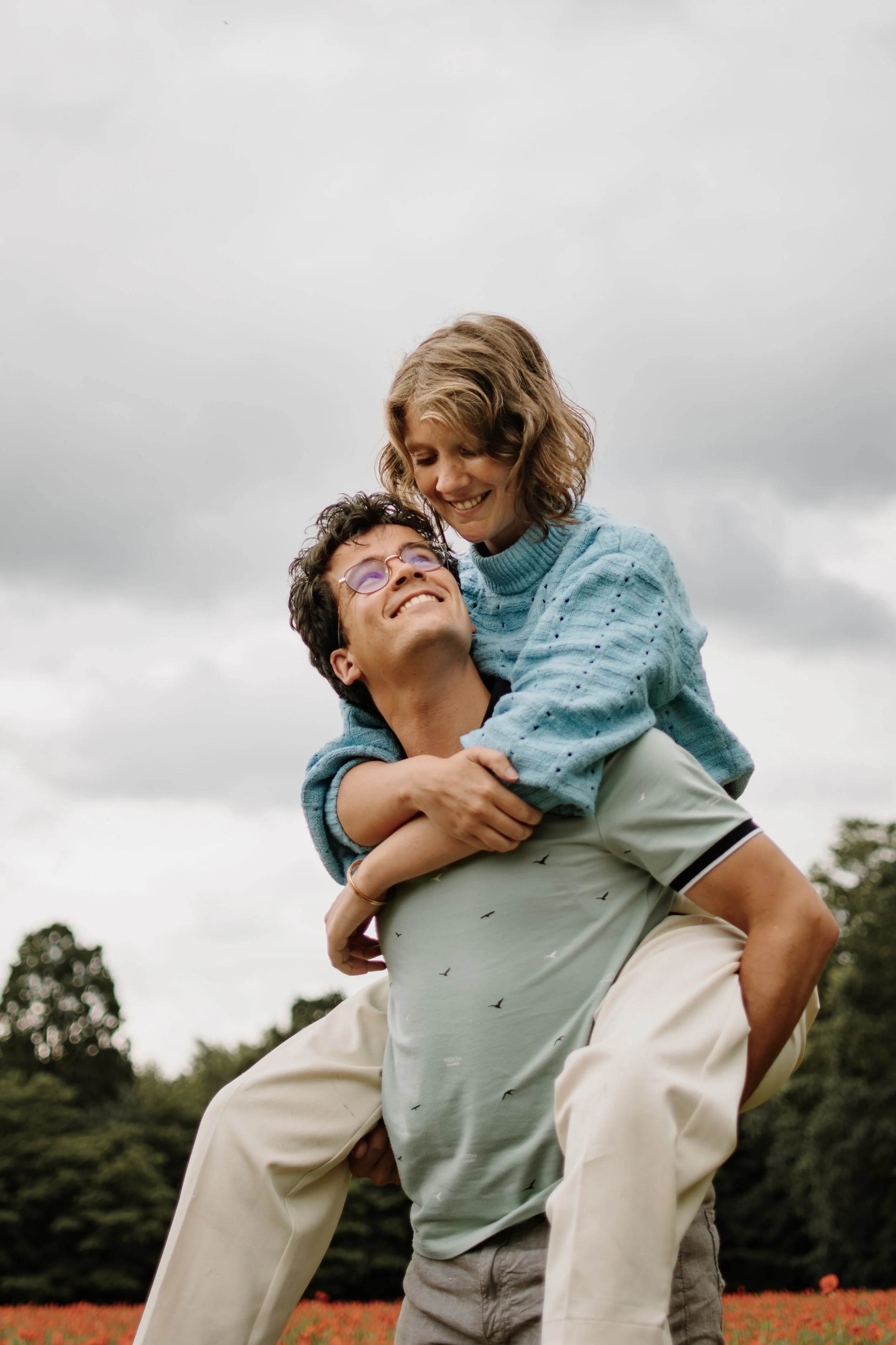 Een man tilt een vrouw op haar rug terwijl ze lachen en genieten in een veld met bloemen onder een bewolkte lucht.