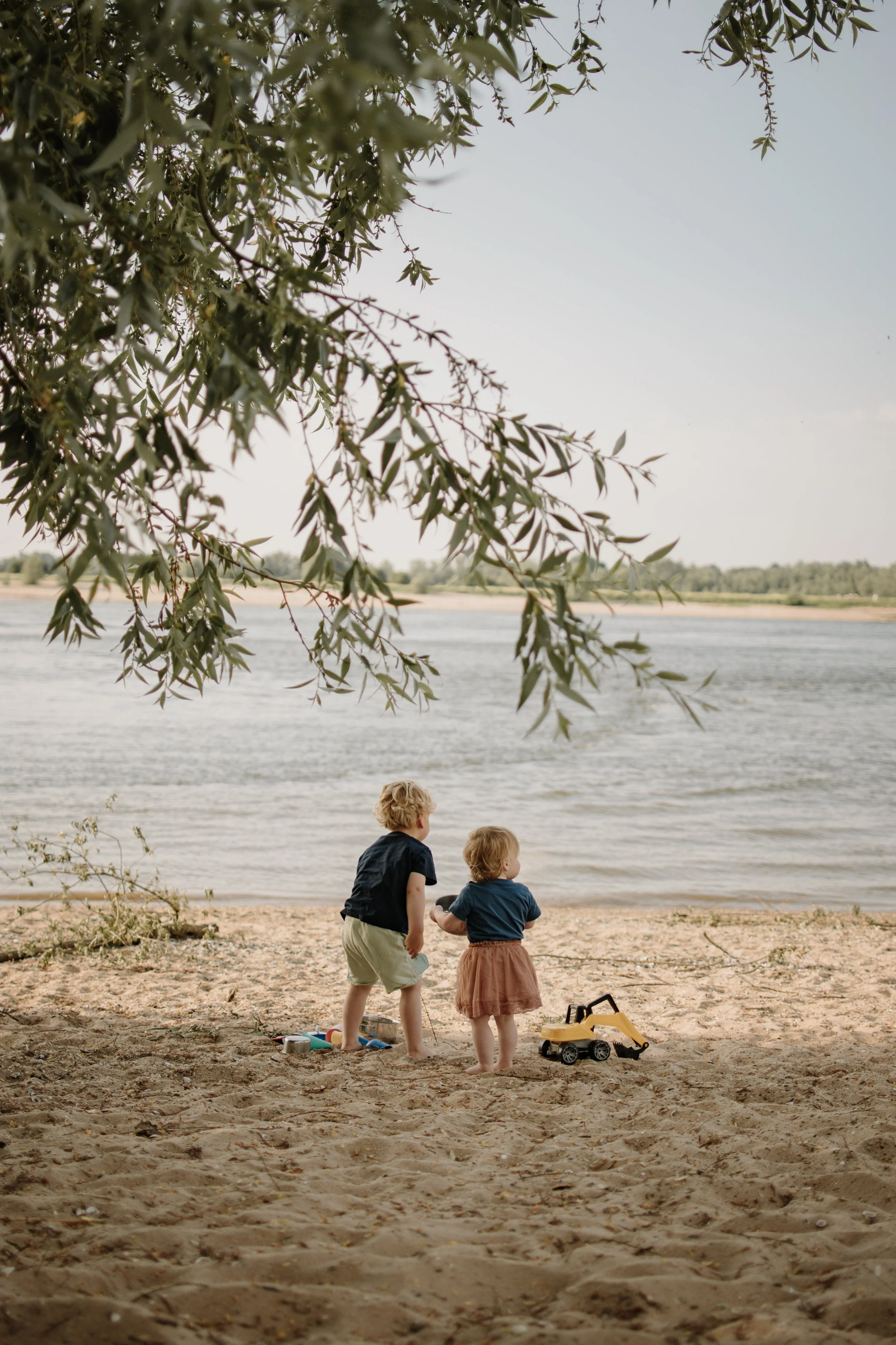 Twee kinderen spelen op het strand bij een rivier, met speelgoedauto's en zand, onder een boom, tijdens een zonnige dag.