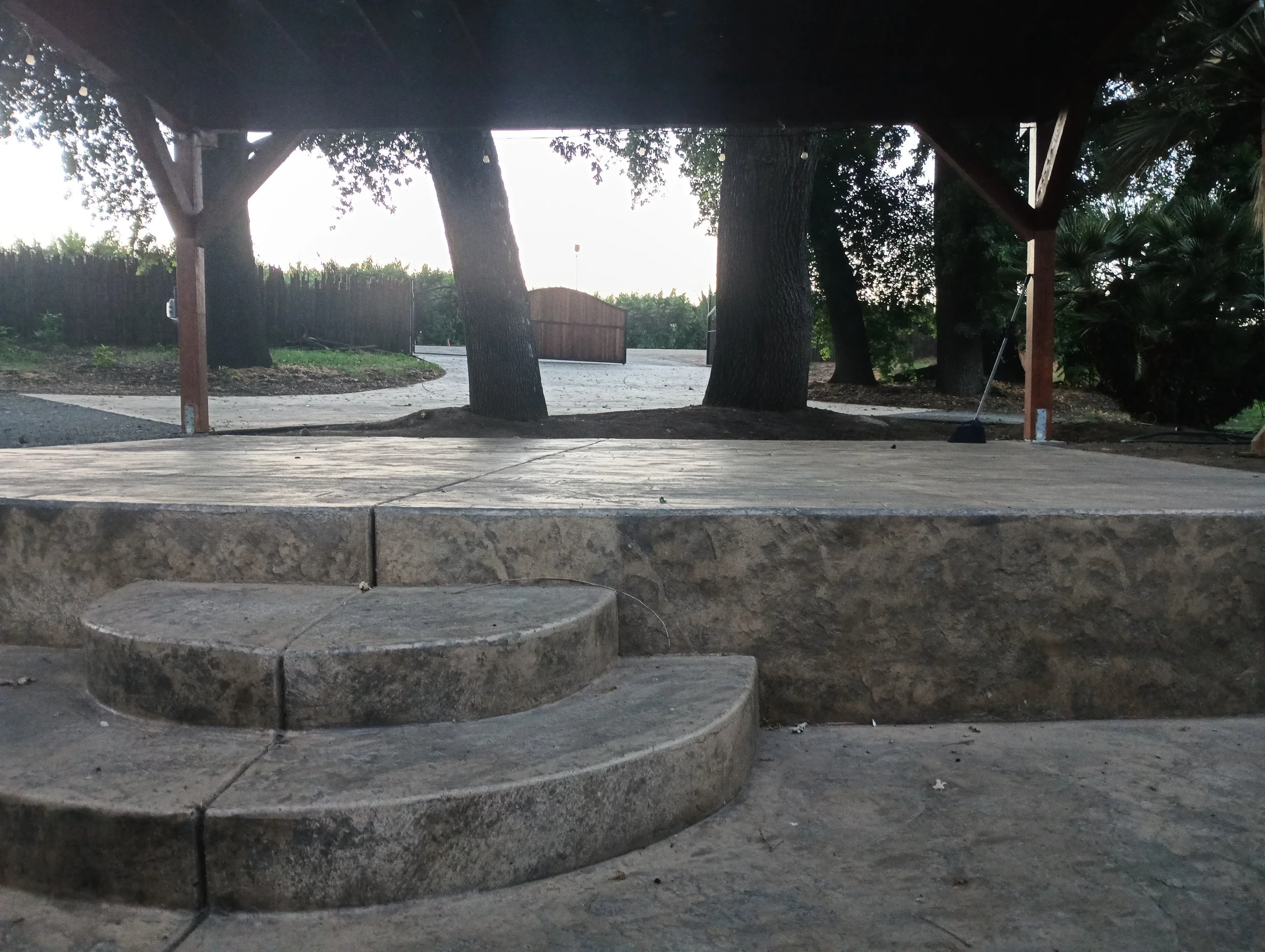 A patio with a stone ledge and steps, underneath a wooden shelter supported by wooden beams. Behind the shelter, there are trees and a gate.