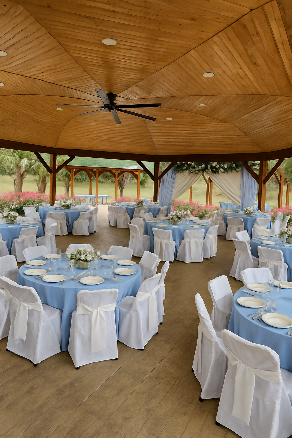 Indoor event space decorated for a formal gathering with round tables covered in blue tablecloths, white chairs with white covers and bows, floral centerpieces, and an open view of outdoors with trees and pink flowers.