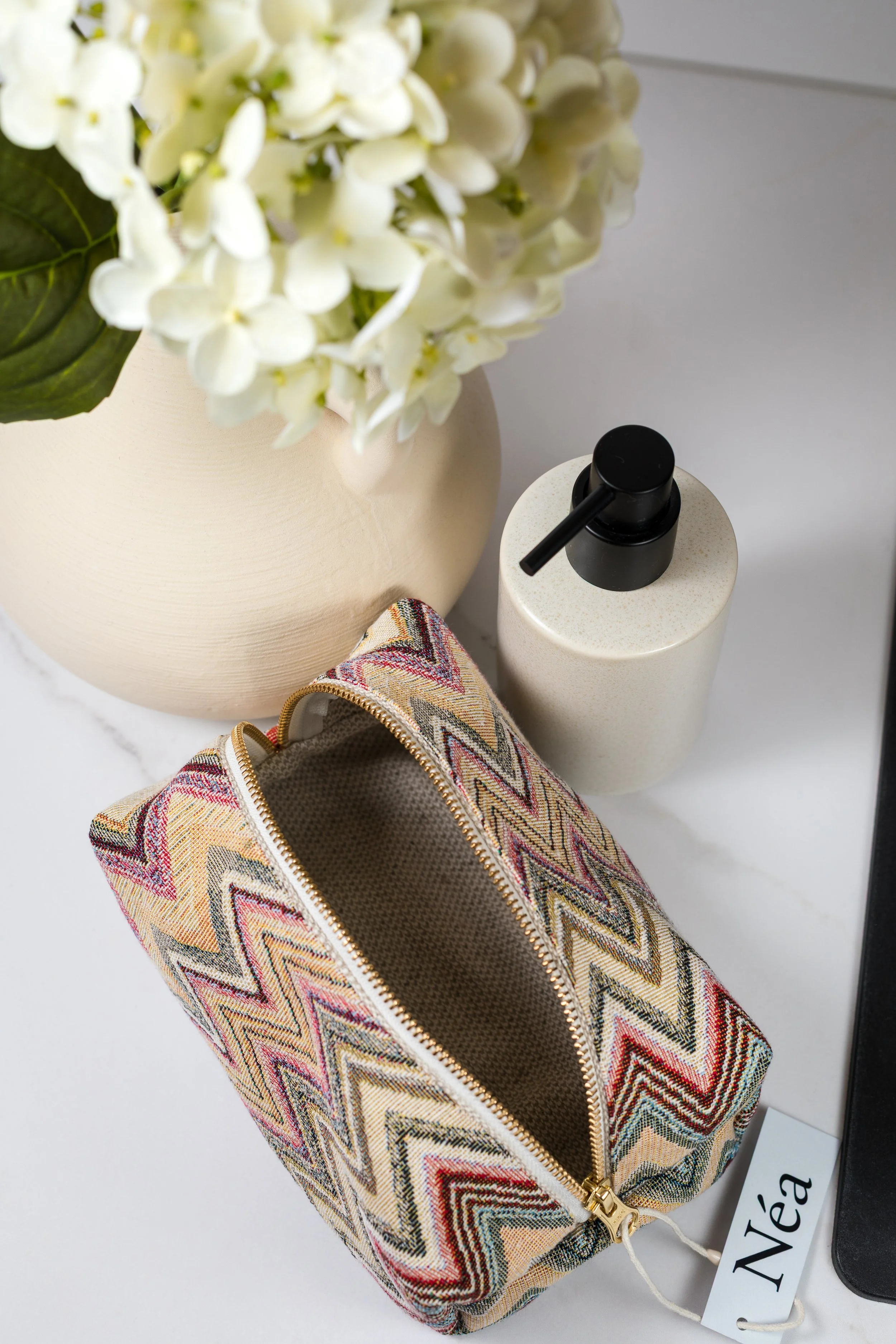 En beige vase med hvite blomster, en hvit væske dispenser med svart pumpe, og en fargerikt mønstret kosmetikkveske med gullfarget glidelås. Et navnmerke merket 'Néa' ligger ved vesken.
