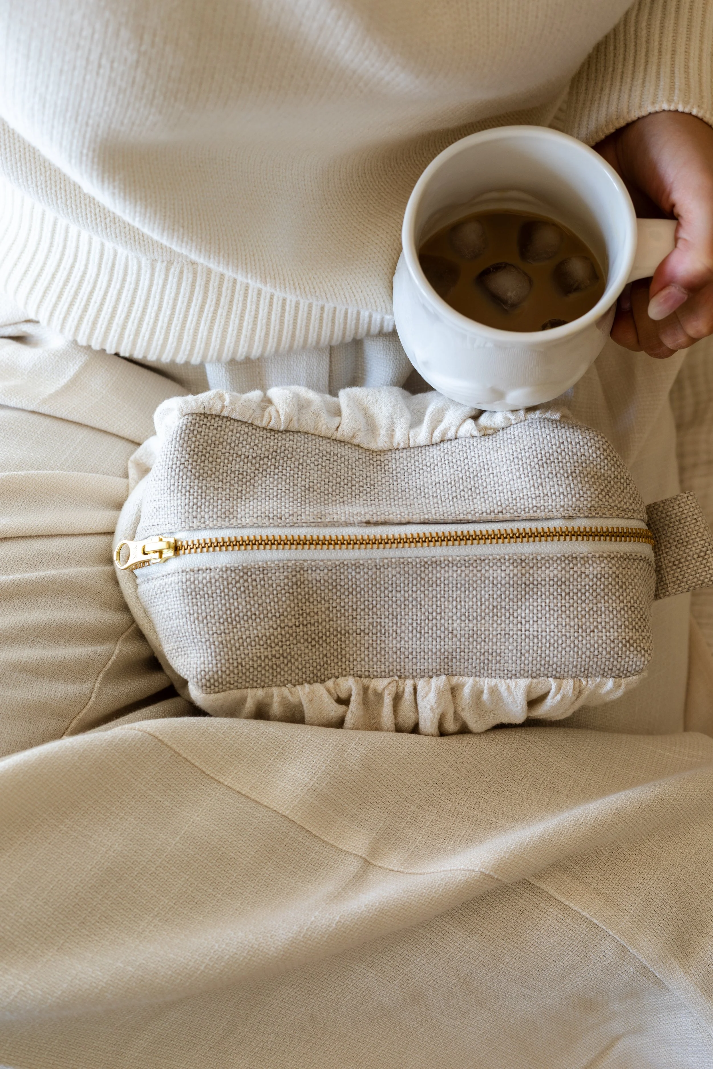 Person holder en kopp med kaffe og isbiter, sittende på et beige stoffsete med en lysgrå liten veske foran.