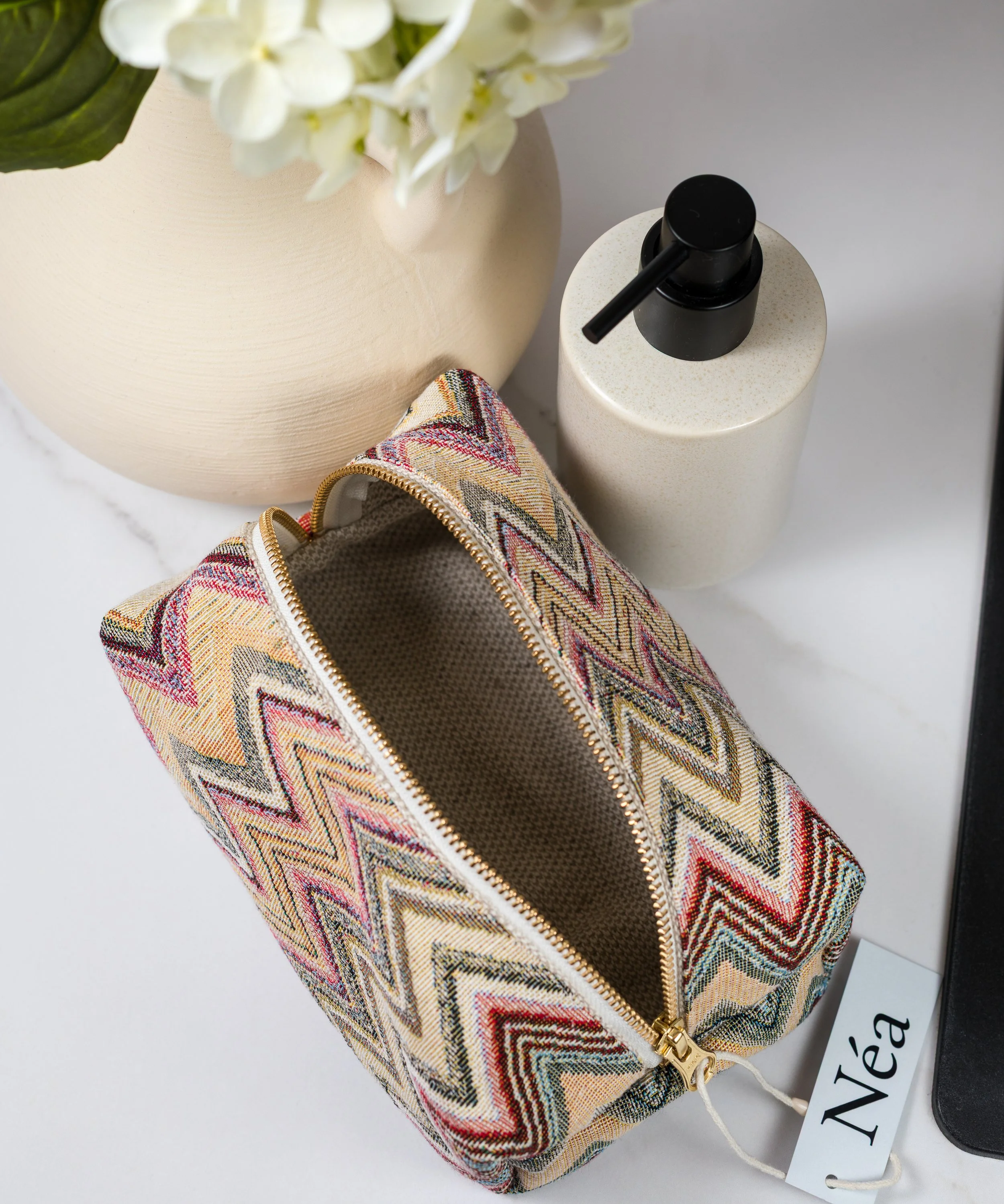 En liten veske med zigzag-mønster i ulike farger, en beige vase med hvite blomster, og en pepperkvern i beige med svart dispenser er plassert på en hvit overflate.