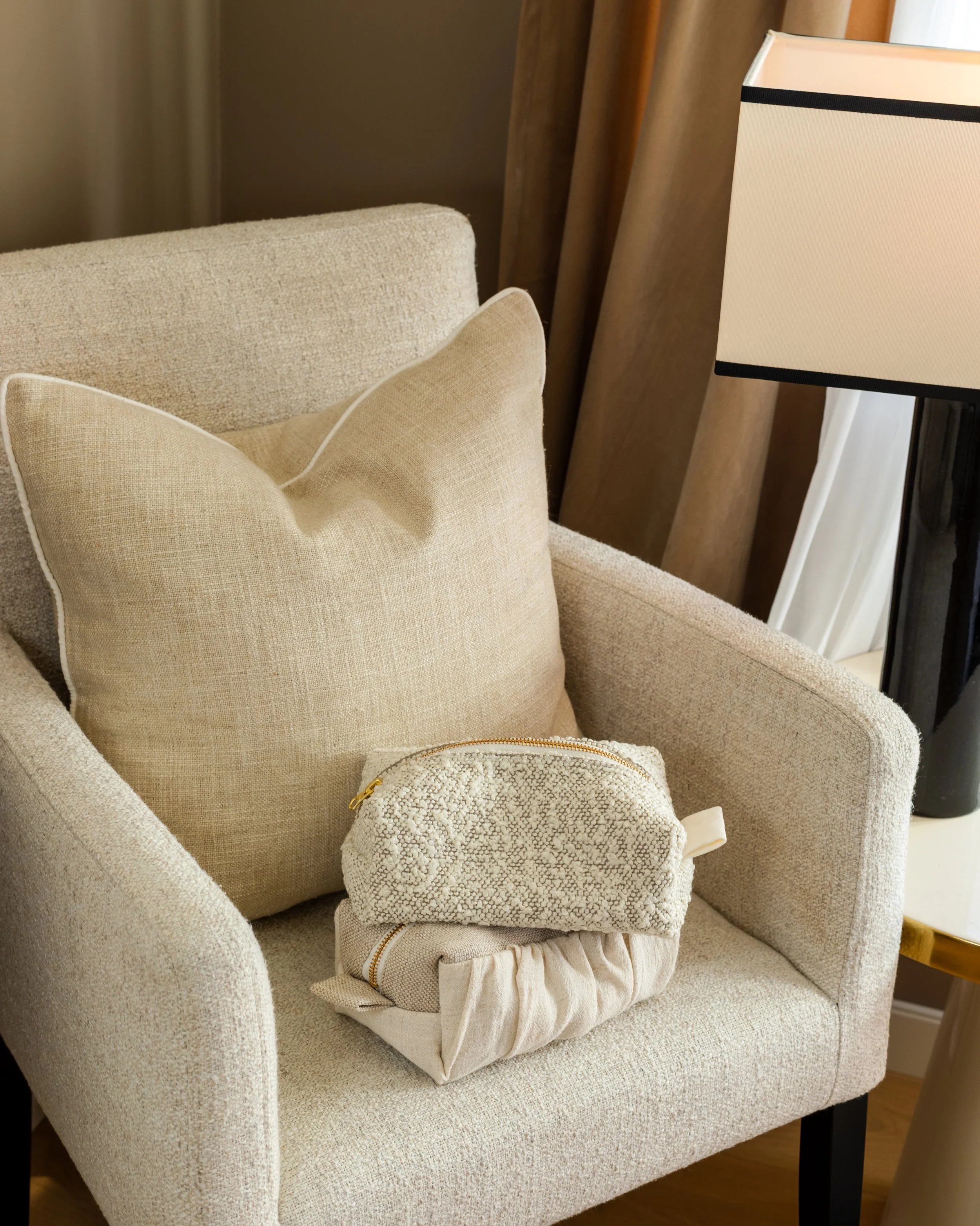 En beige lenestol med en beige pute og en liten, hvit og mønstret veske på. En stokkelys i svart og hvitt står ved siden av, med en del av vindu med hvite gardiner i bakgrunnen.