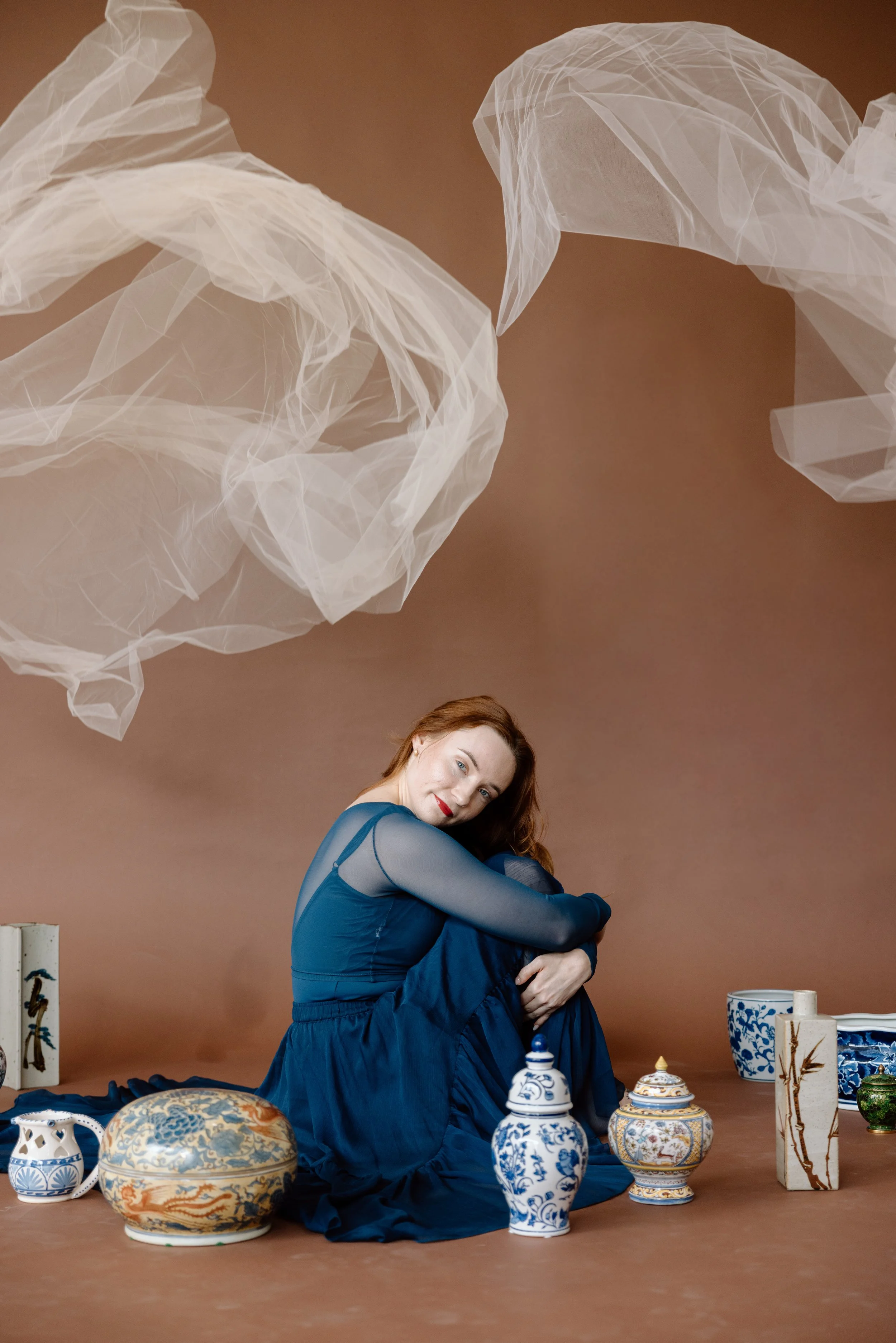 A woman in a blue dress sitting on the ground surrounded by decorative porcelain vases and ceramics, with large white fabric or tulle hanging overhead against a brown background.