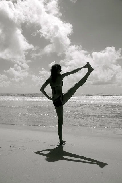 A woman performing a yoga pose on a beach, holding her leg high with one hand, with clouds in the sky.