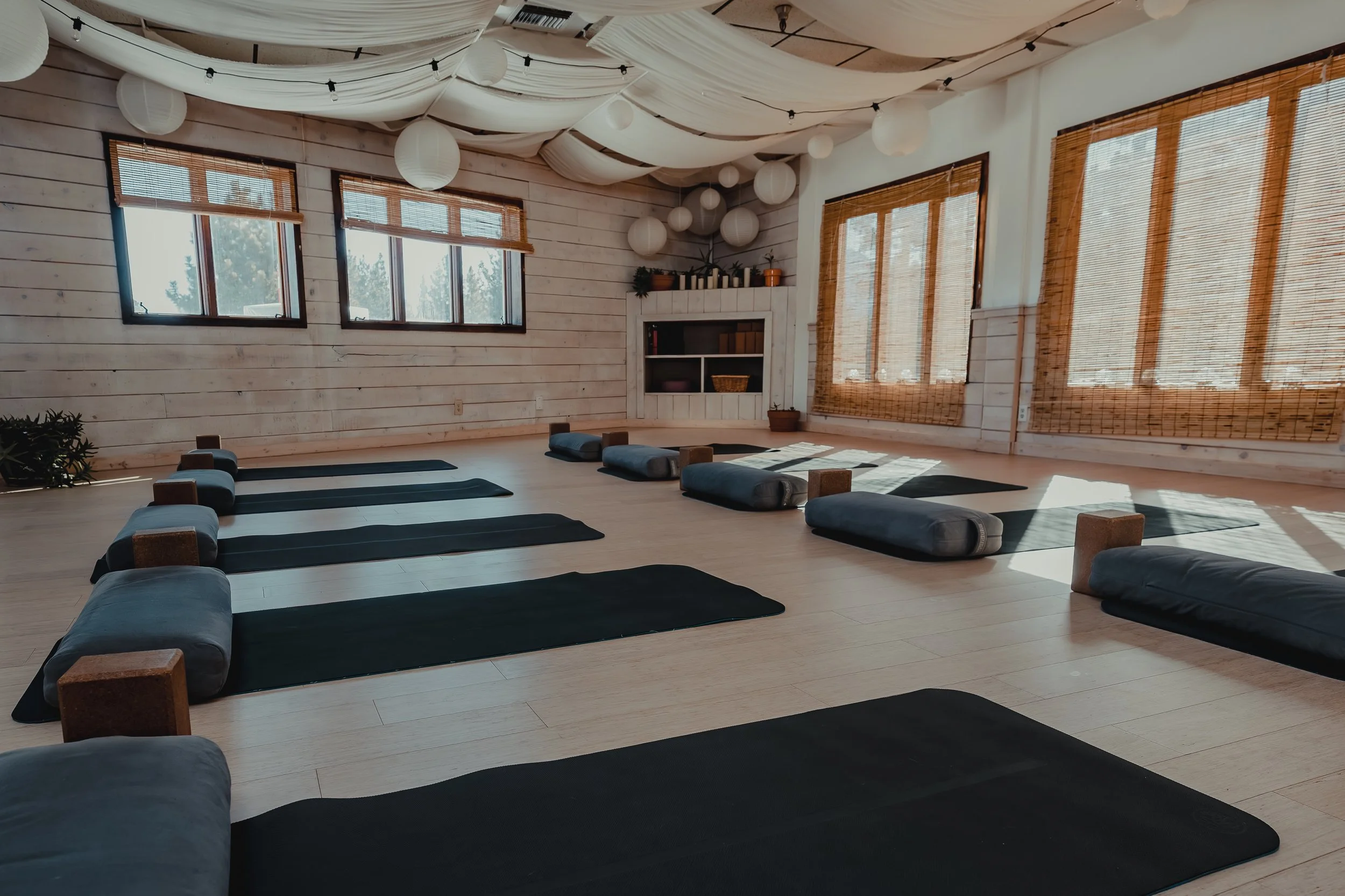 A yoga or meditation studio with black mats and bolsters on light wooden floors, decorated with paper lanterns and draped fabric on the ceiling, large windows with bamboo blinds, and a white shelf with plants and decor.