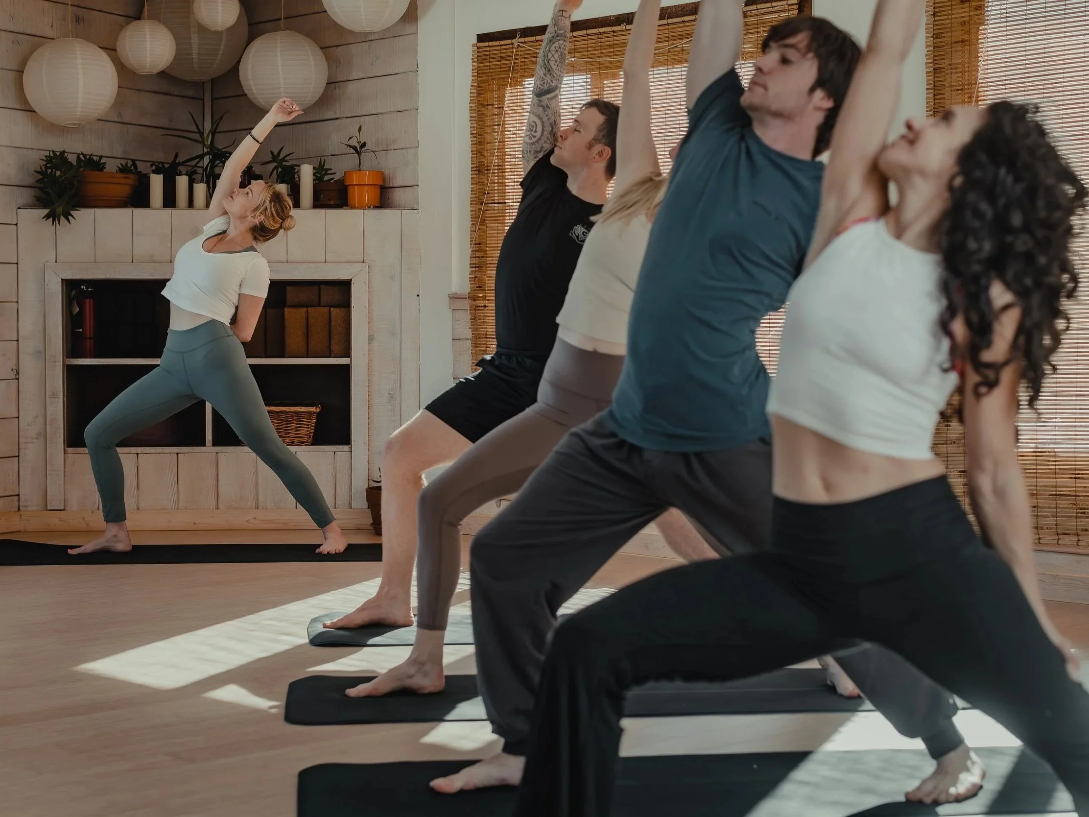 A woman leading a yoga class with four participants in a room with wooden decor and hanging paper lanterns, all doing a side stretch pose on mats.