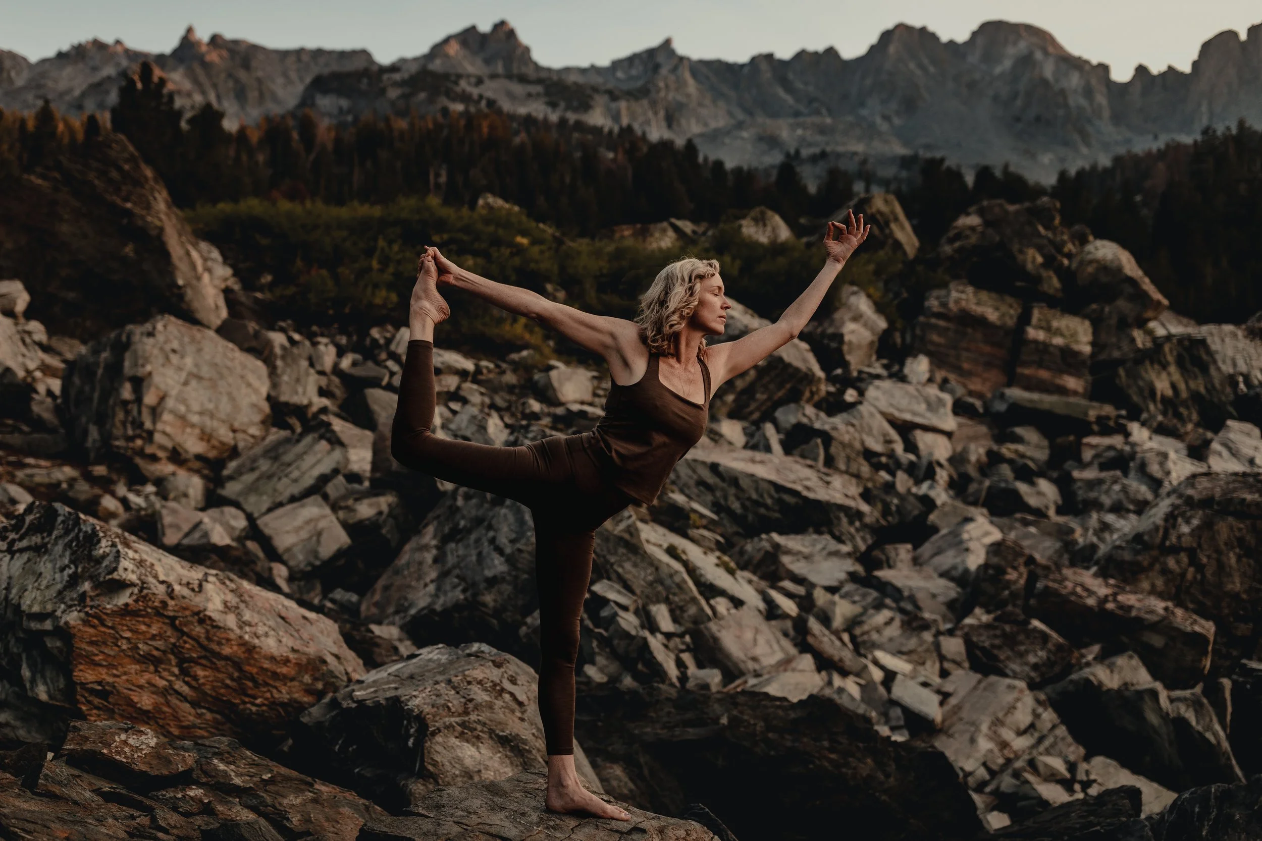 A woman practicing yoga or dance outdoors on a rocky terrain, performing a one-legged balancing pose with a mountain range in the background during sunset or sunrise.