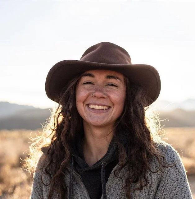 Young woman with long curly hair, smiling, wearing a broad-brimmed hat and a fleece jacket outdoors in sunlight.