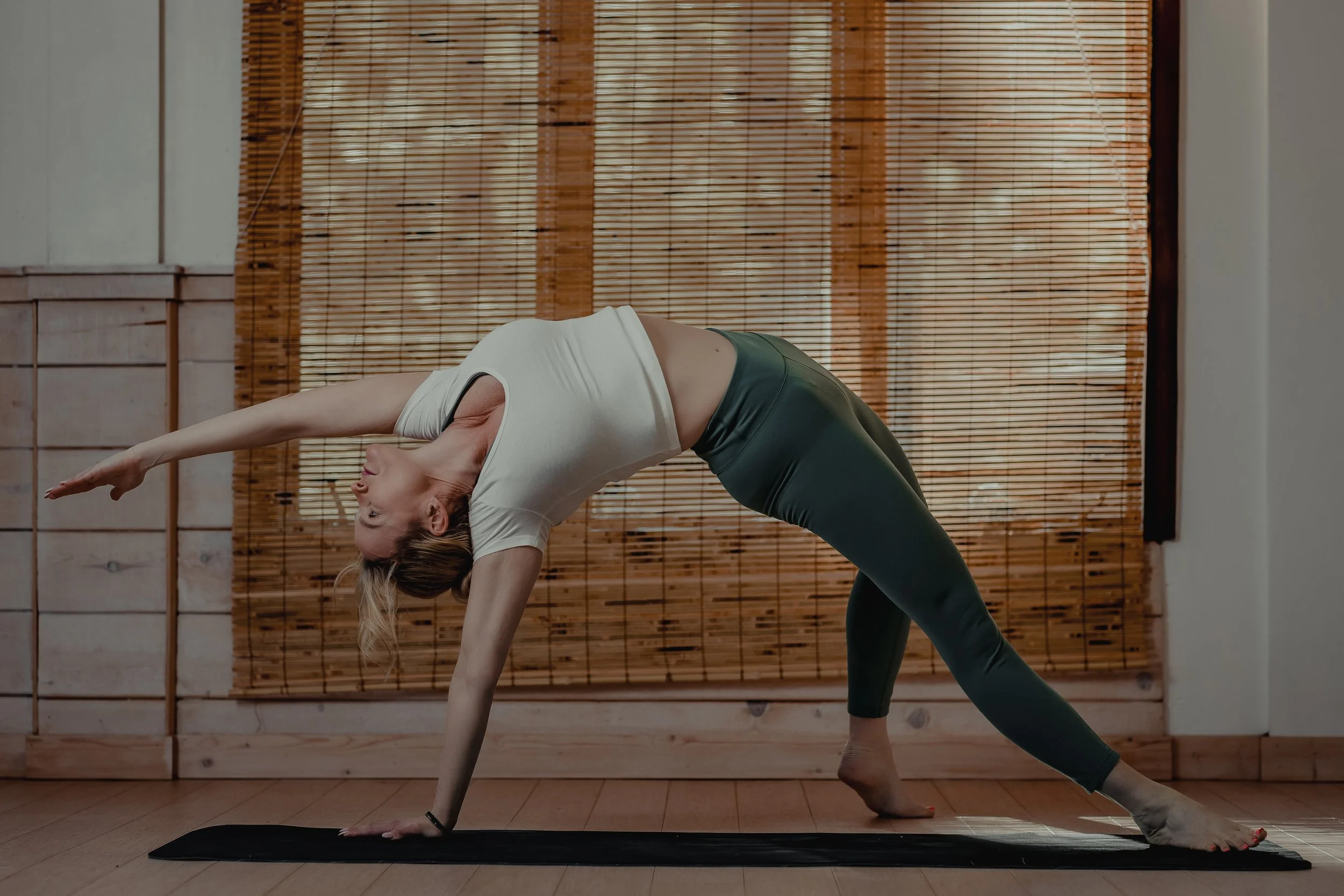 A woman practicing yoga indoors on a black mat, performing a downward-facing dog pose with her head turned to the side, against a background of bamboo blinds and wooden paneling.
