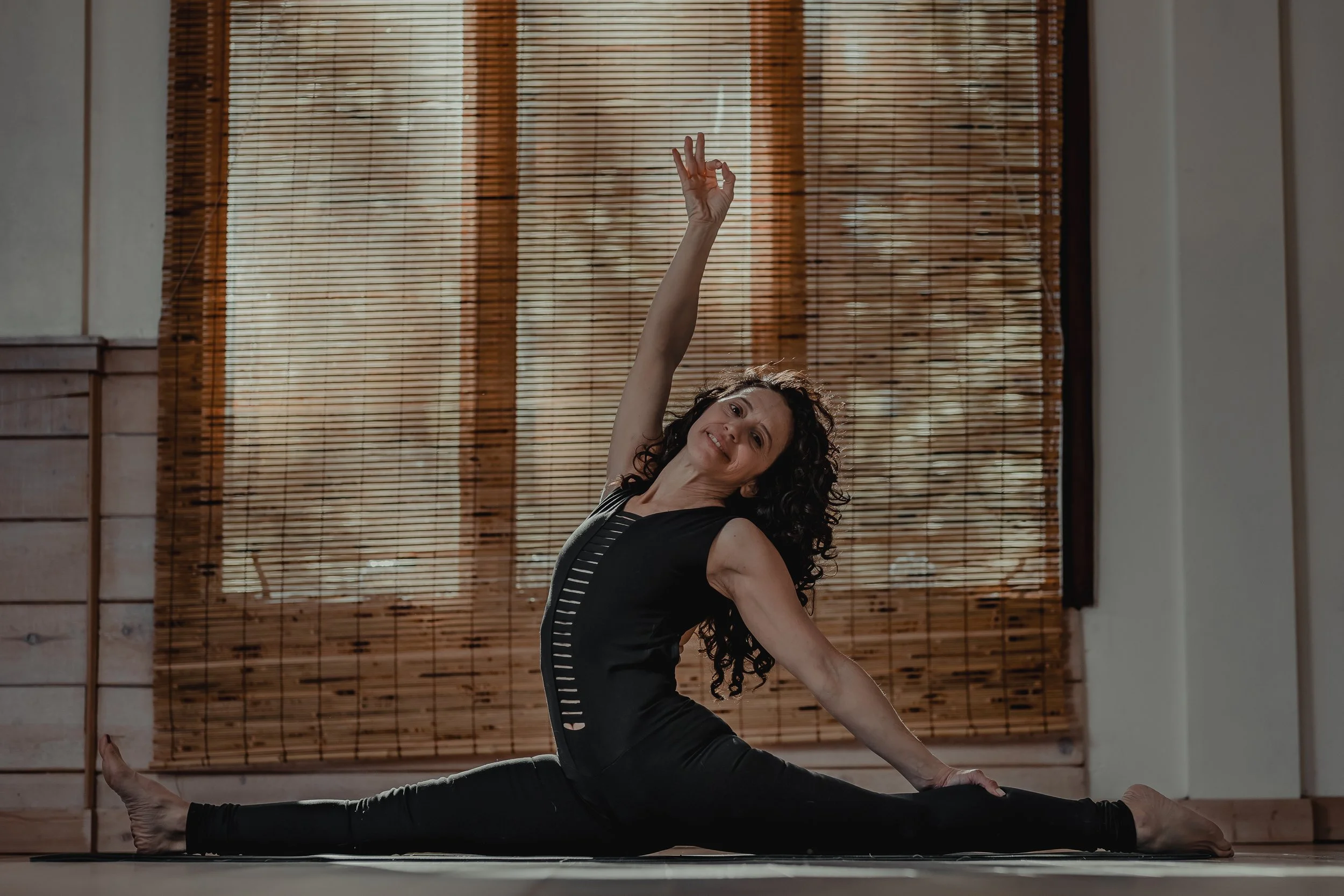 Woman doing a seated split yoga pose indoors with bamboo blinds in the background.