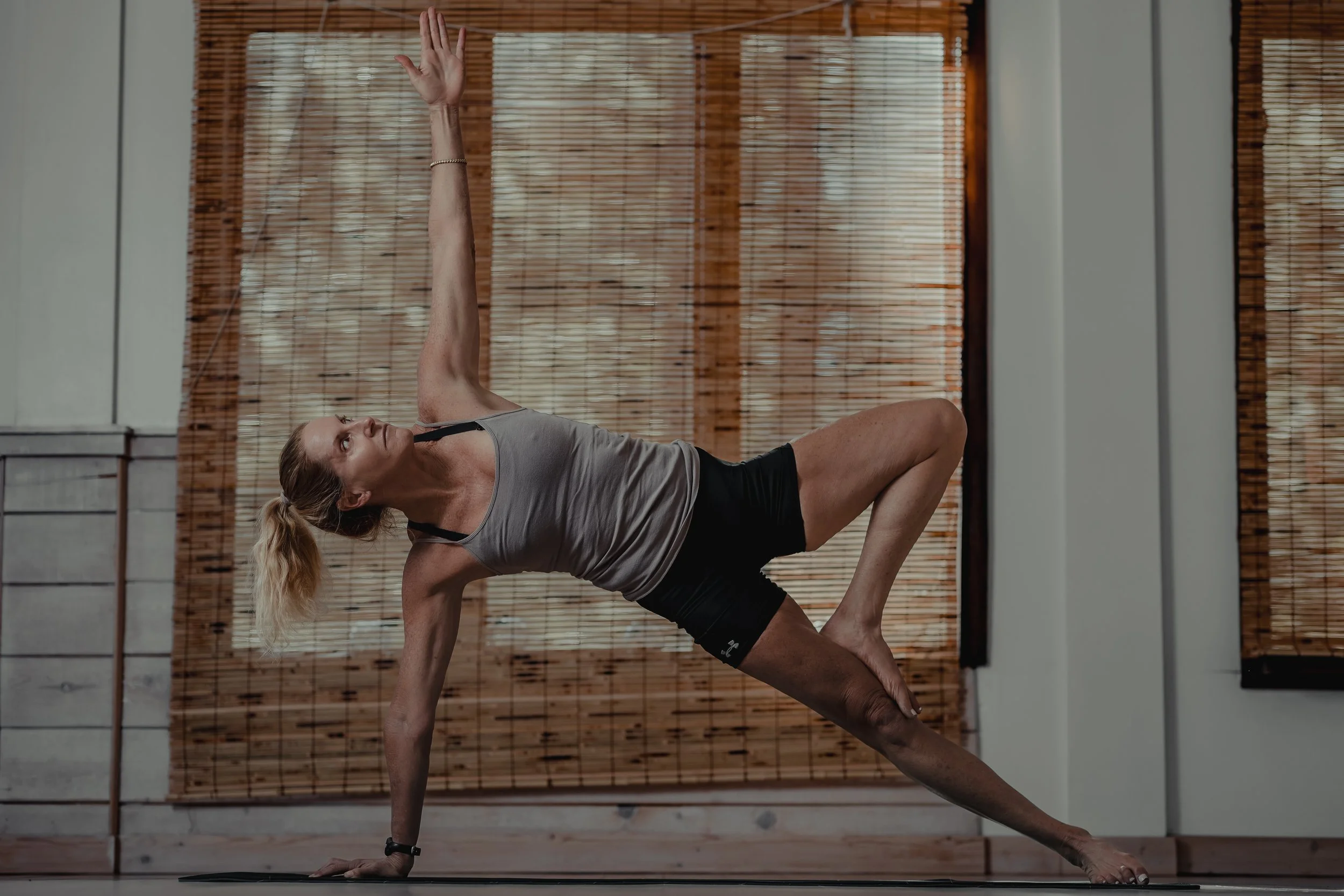 A woman practicing yoga indoors, balancing on one hand and foot in a pose with her other hand raised and looking upwards.