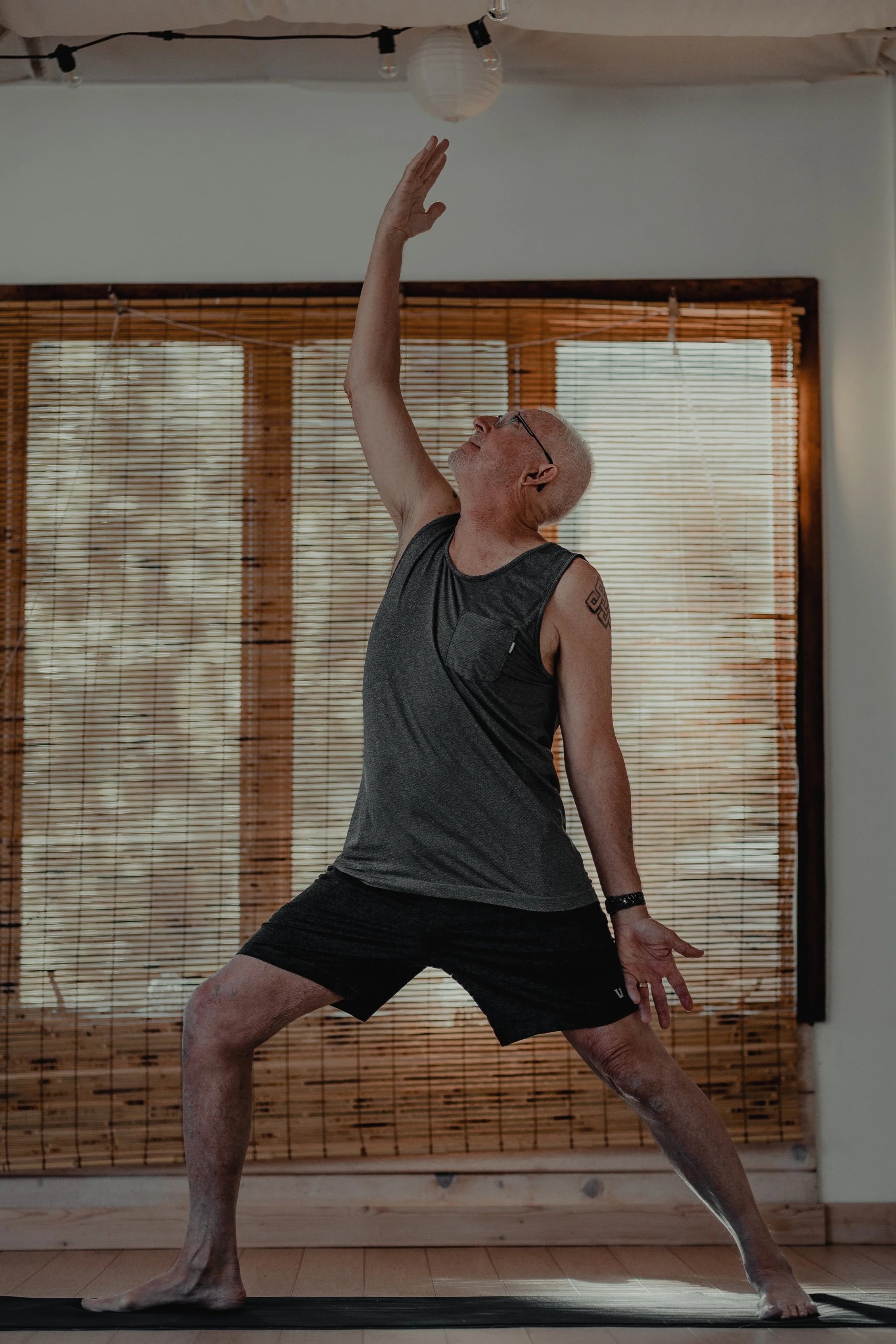 An elderly man with glasses practicing yoga indoors, standing in a wide-legged stance with one arm extended upward and the other extended behind him, on a black yoga mat.