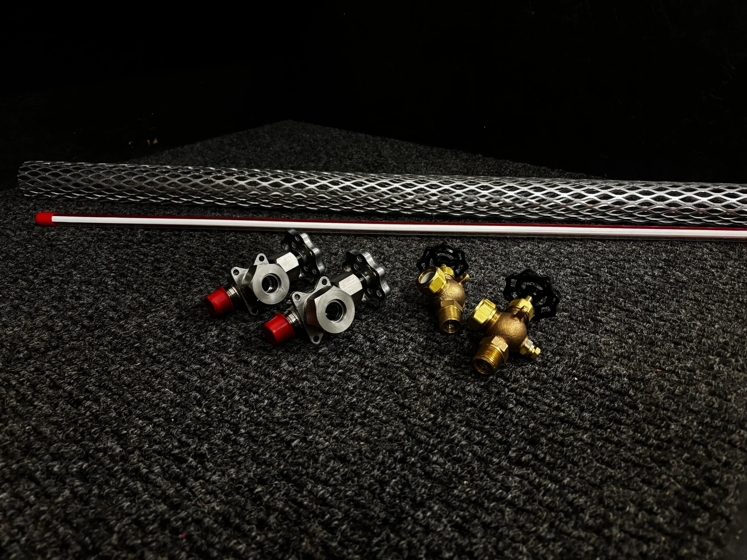 Two silver metal water pump fittings with red caps, two brass water pump fittings with black valve handles, a metal mesh pipe guard, and two long metal rods with red tips on a black textured surface.