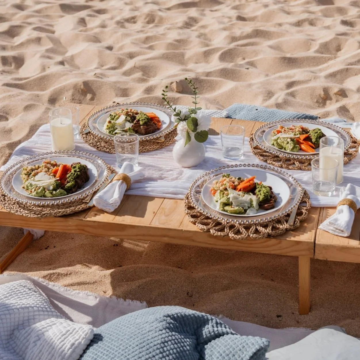 Plated dinners on woven chargers at a low table set up in the sand for a beach event