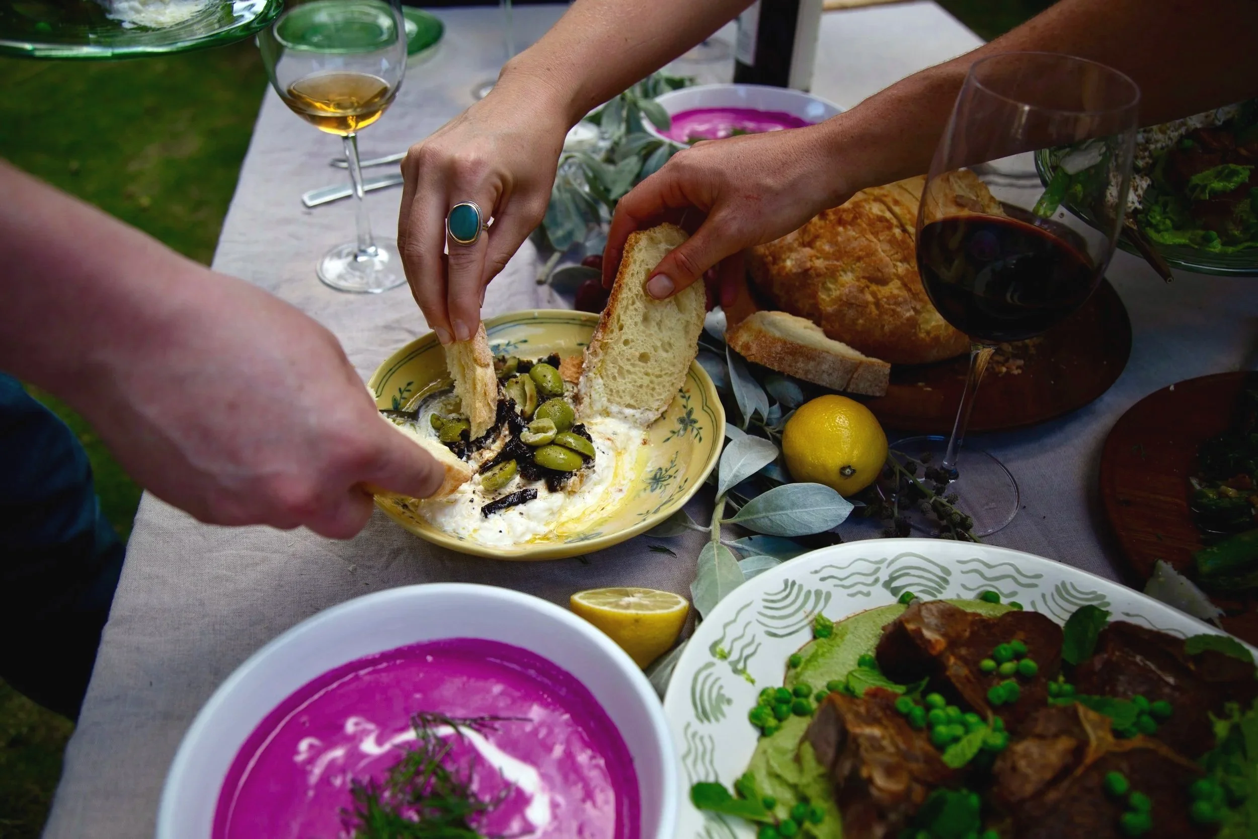 Guests tearing bread over marinated olives and stracciatella at a family-style private dinner with chilled beet soup and wine