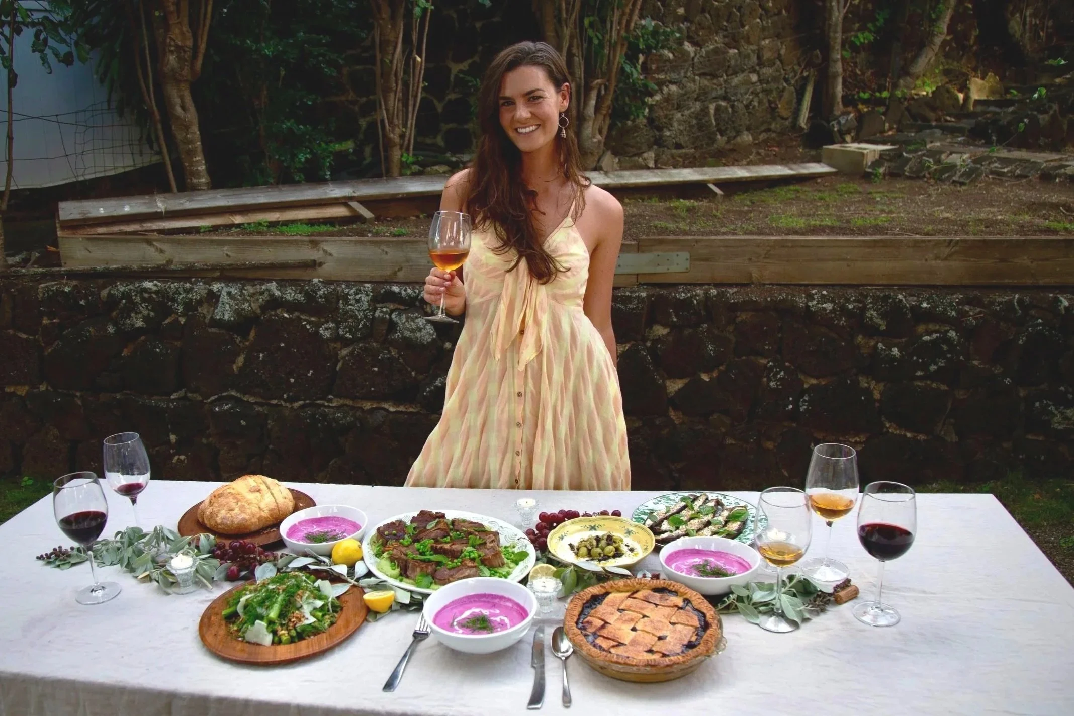 Chef Carli Parisi holding a glass of wine beside a long outdoor table set with breads, salads, chilled beet soup and a pie