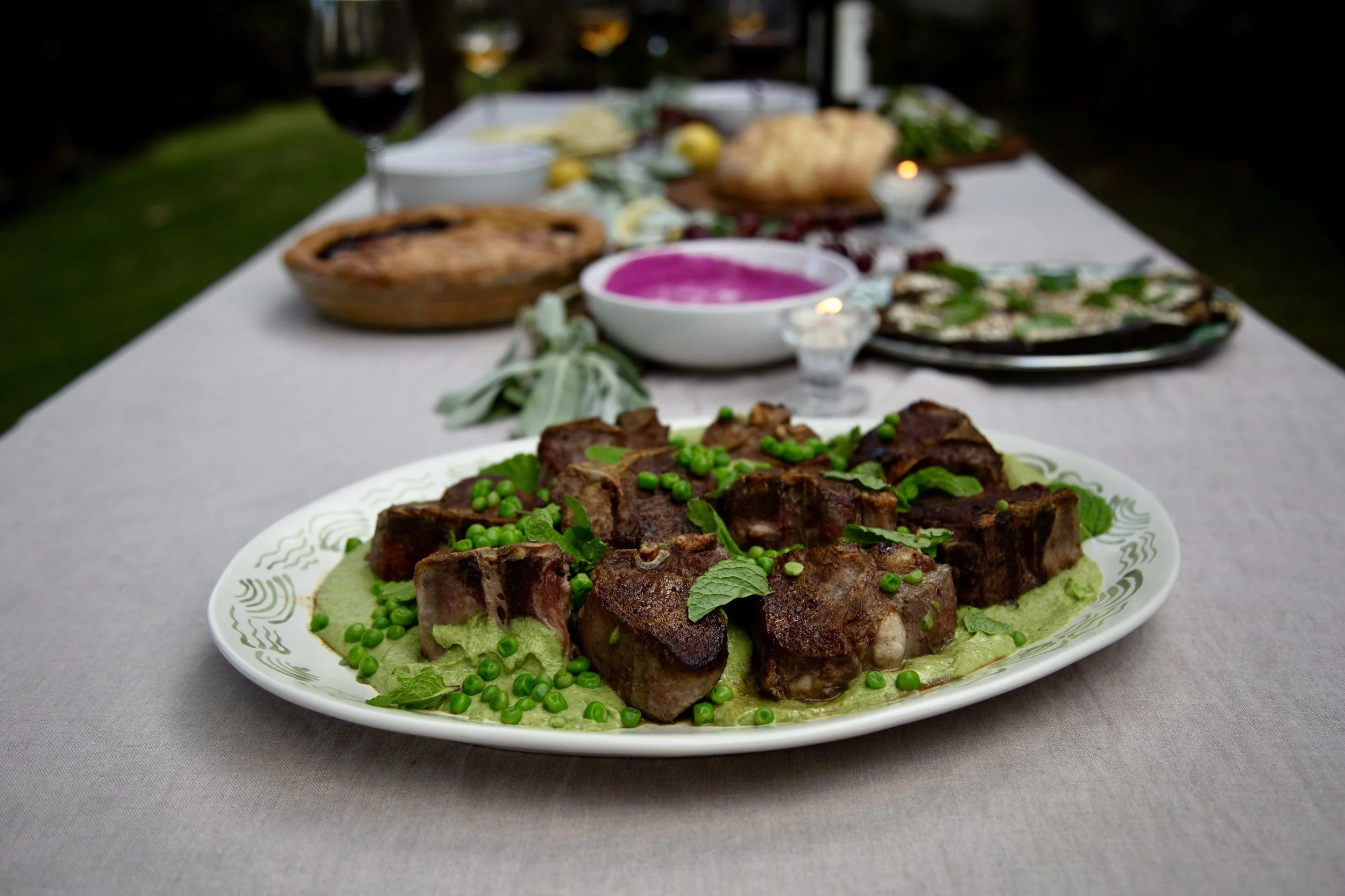 Seared lamb with spring peas and fresh mint on a platter, with a long dinner table set behind it