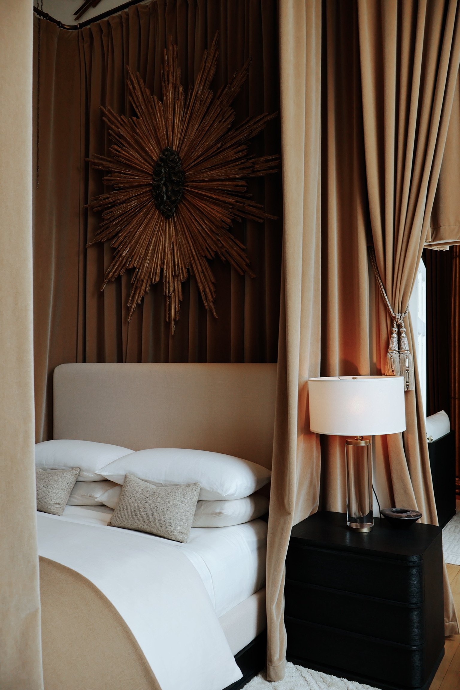 Luxurious hotel bedroom with white bed, beige pillows, dark nightstand, modern lamp, and decorative wooden wall art, surrounded by beige curtains.