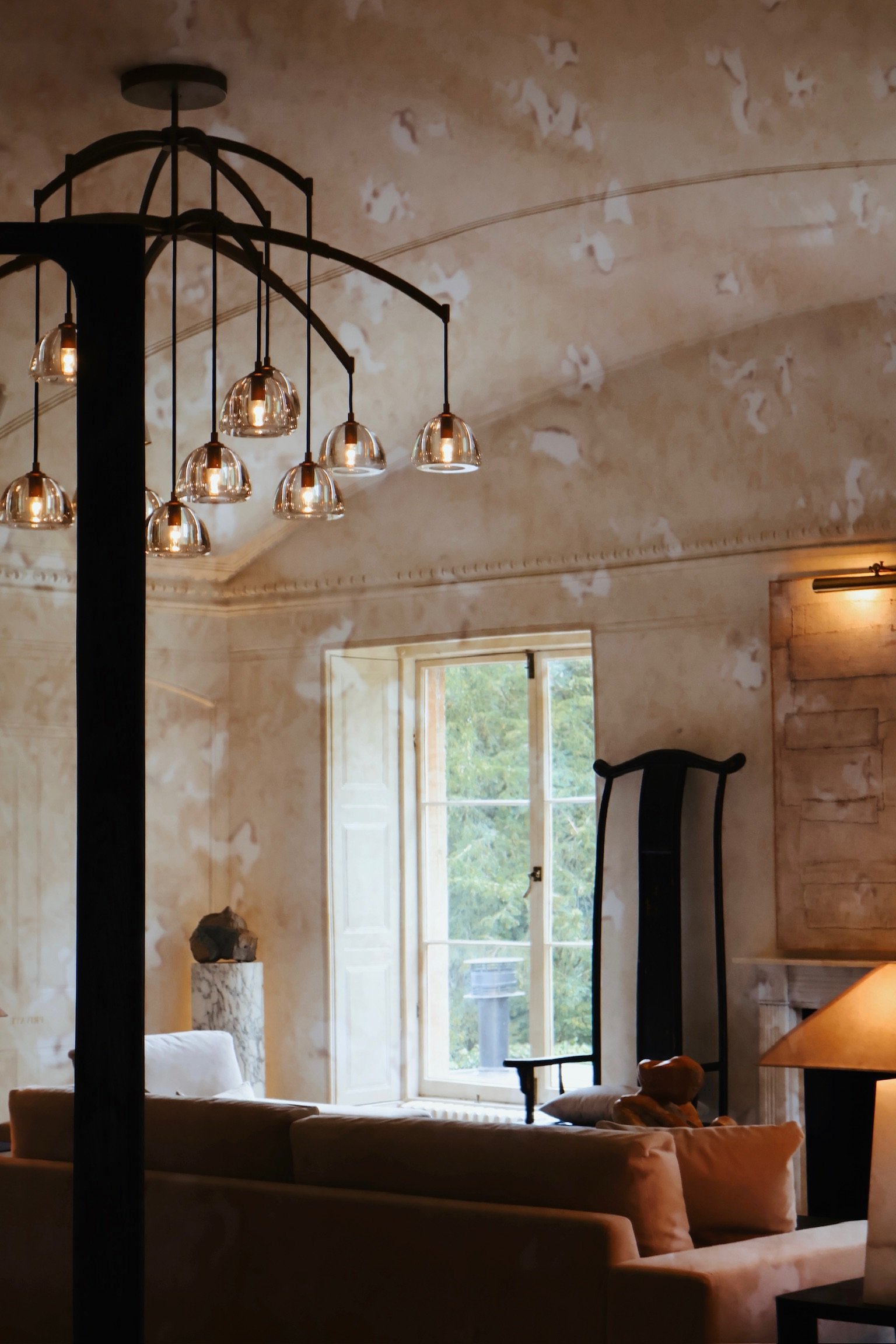 Interior of a cozy living room with a cream-colored sofa, a large window showing greenery outside, a modern chandelier with multiple glass bulbs, and a tall black mirror leaning against the wall.
