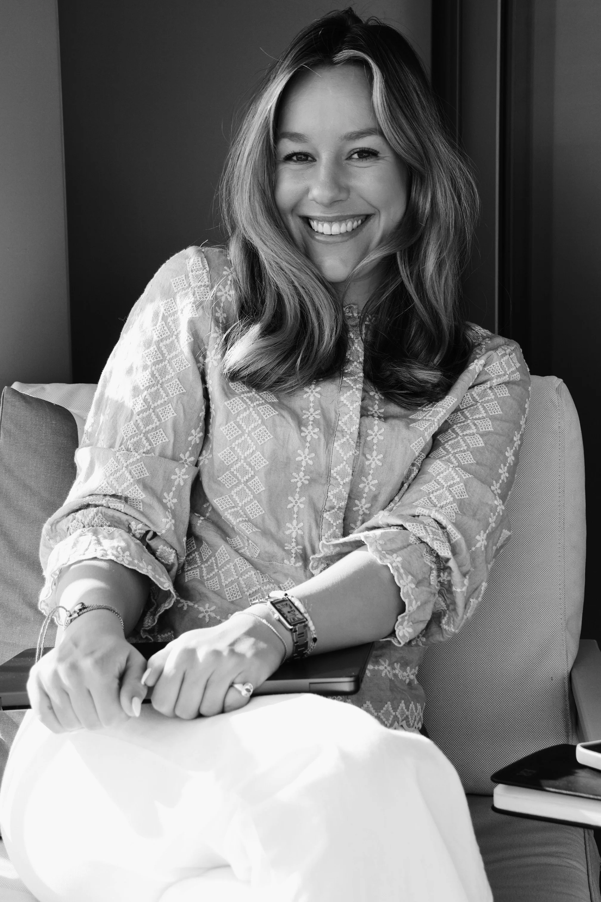 A woman smiling and sitting on a chair, holding a closed MacBook laptop in her lap, with a smartphone and notebook on her side.
