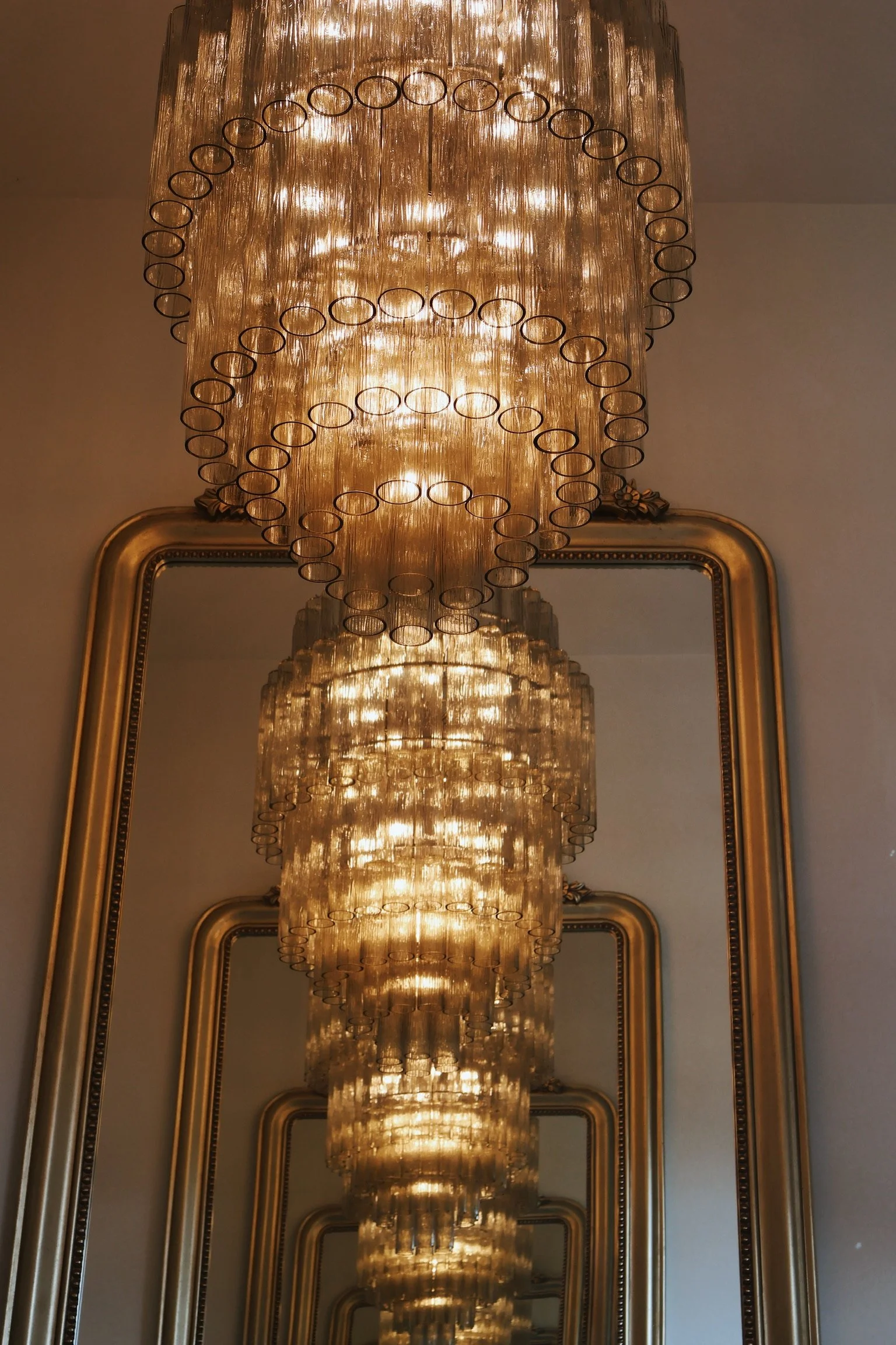 A grand chandelier with multiple tiers of glass and circular metal accents, reflected in a large gold-framed mirror.