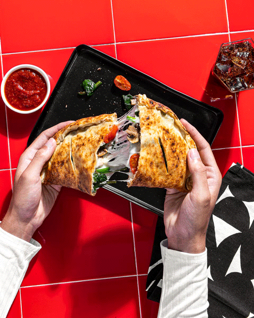 Person holding a slice of stuffed pizza with visible cheese, vegetables, and meats, on a black rectangular plate, with a side of red sauce, a glass of soda, and a black-and-white patterned napkin on a red tiled table.