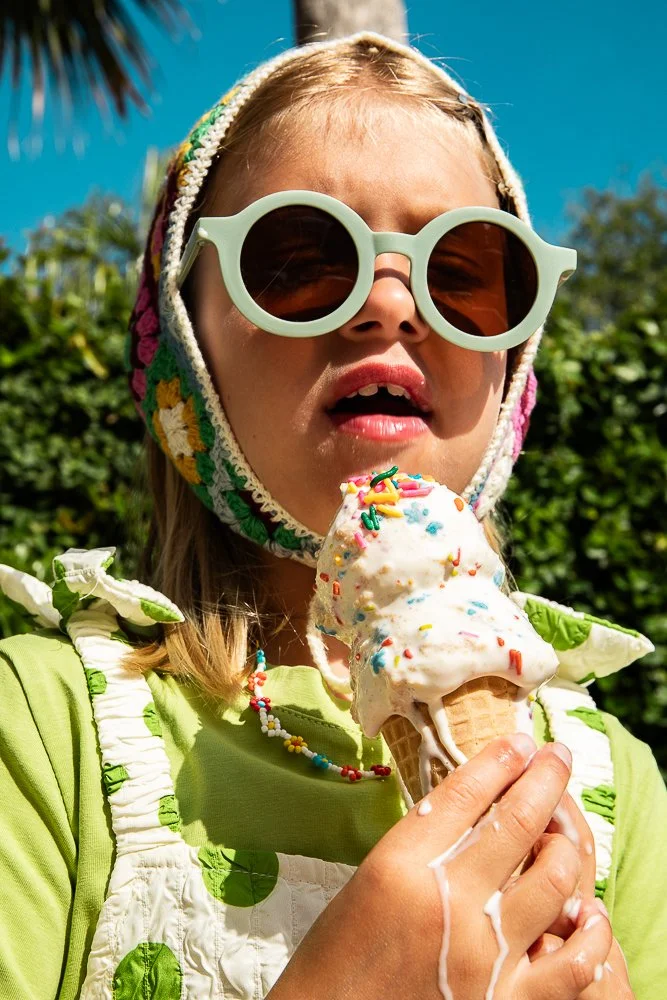 Young girl wearing large sunglasses and a colorful knit hood, holding an ice cream cone with sprinkles, outdoors on a sunny day.