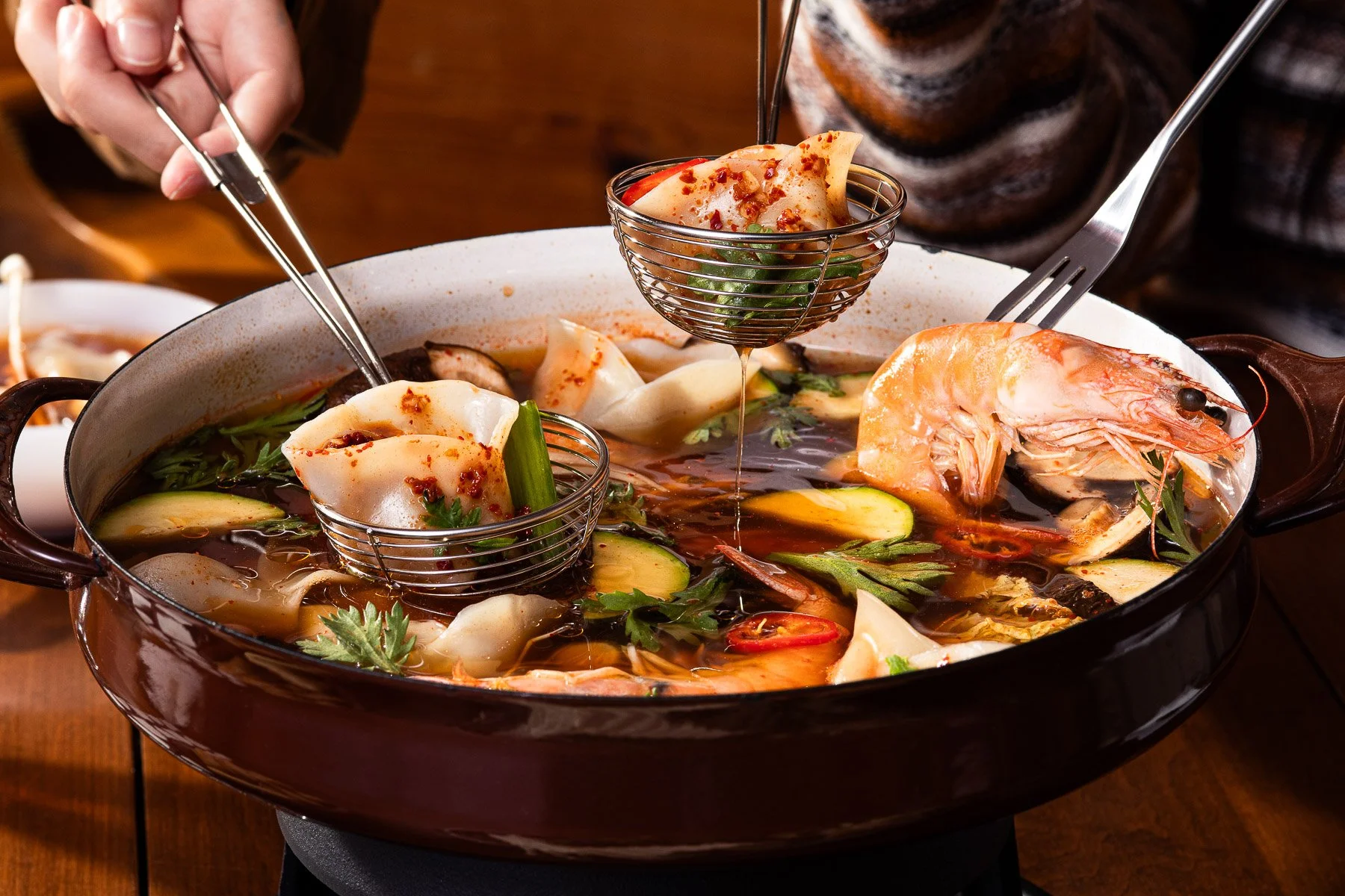 Hot pot with shrimp, vegetables, and dumplings, with a person using tongs to add dumplings into the broth.