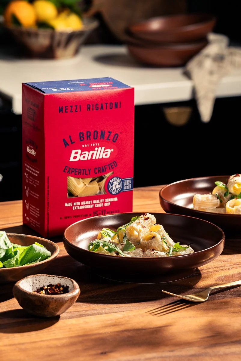 A box of Barilla pasta on a wooden table, with cooked pasta and fresh basil in bowls nearby, and a small dish of red pepper flakes.