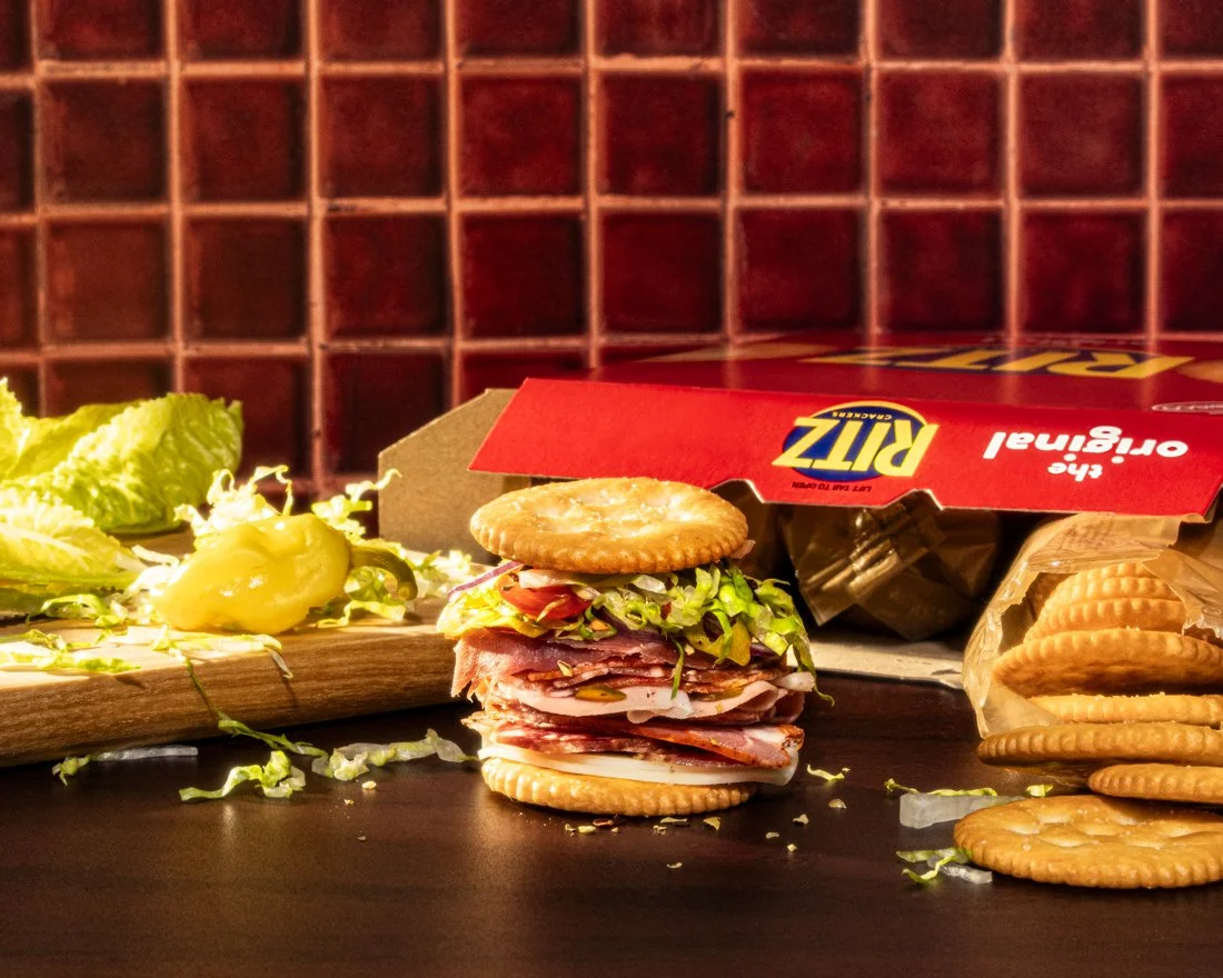 Italian sub sandwich made with crackers, lettuce, tomato, and cold cuts, placed on a dark surface with an open box of Ritz crackers and some lettuce and sliced vegetables in the background.